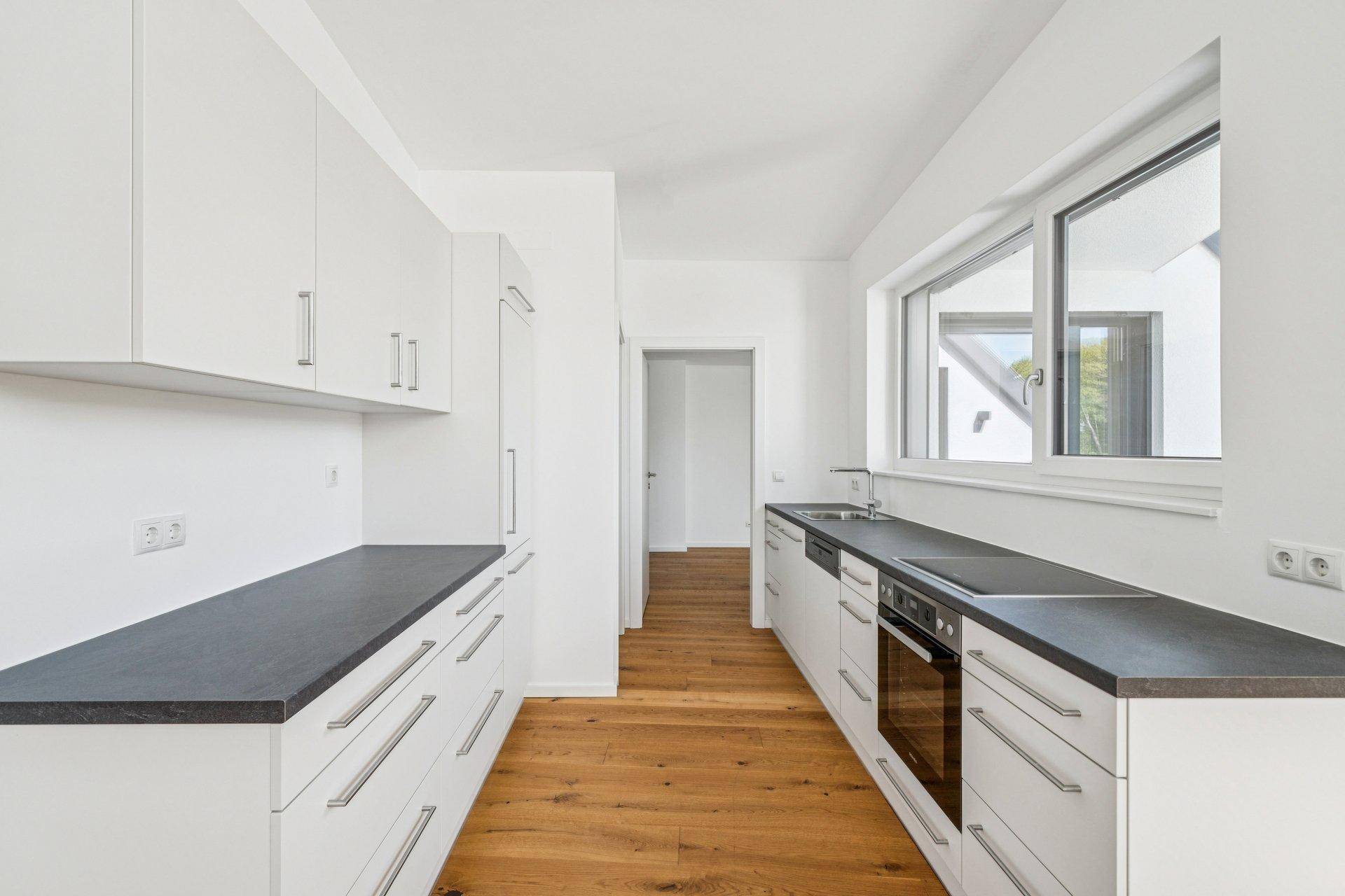 Moderne Küche mit weißen Schränken, dunklen Arbeitsplatten und Holzboden, gut beleuchtet durch ein Fenster.