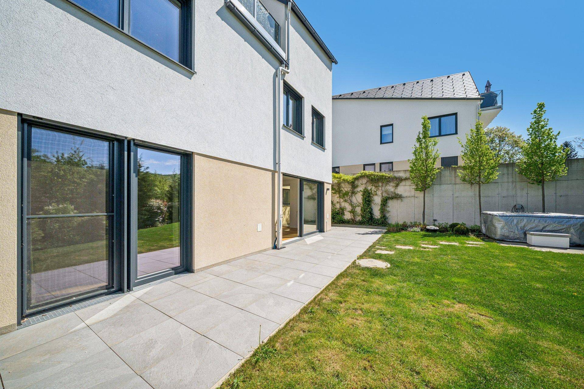 Moderne Hausfassade mit großen Fenstern und Zugang zum gepflegten Garten mit Whirlpool.