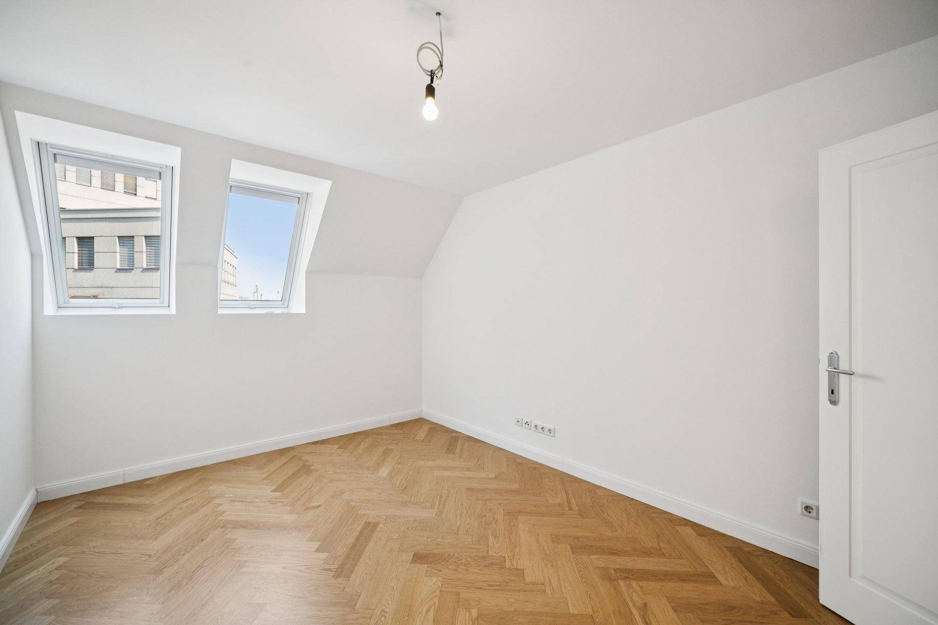Leerer Raum mit Dachschrägen, Parkettboden und zwei Dachfenstern.