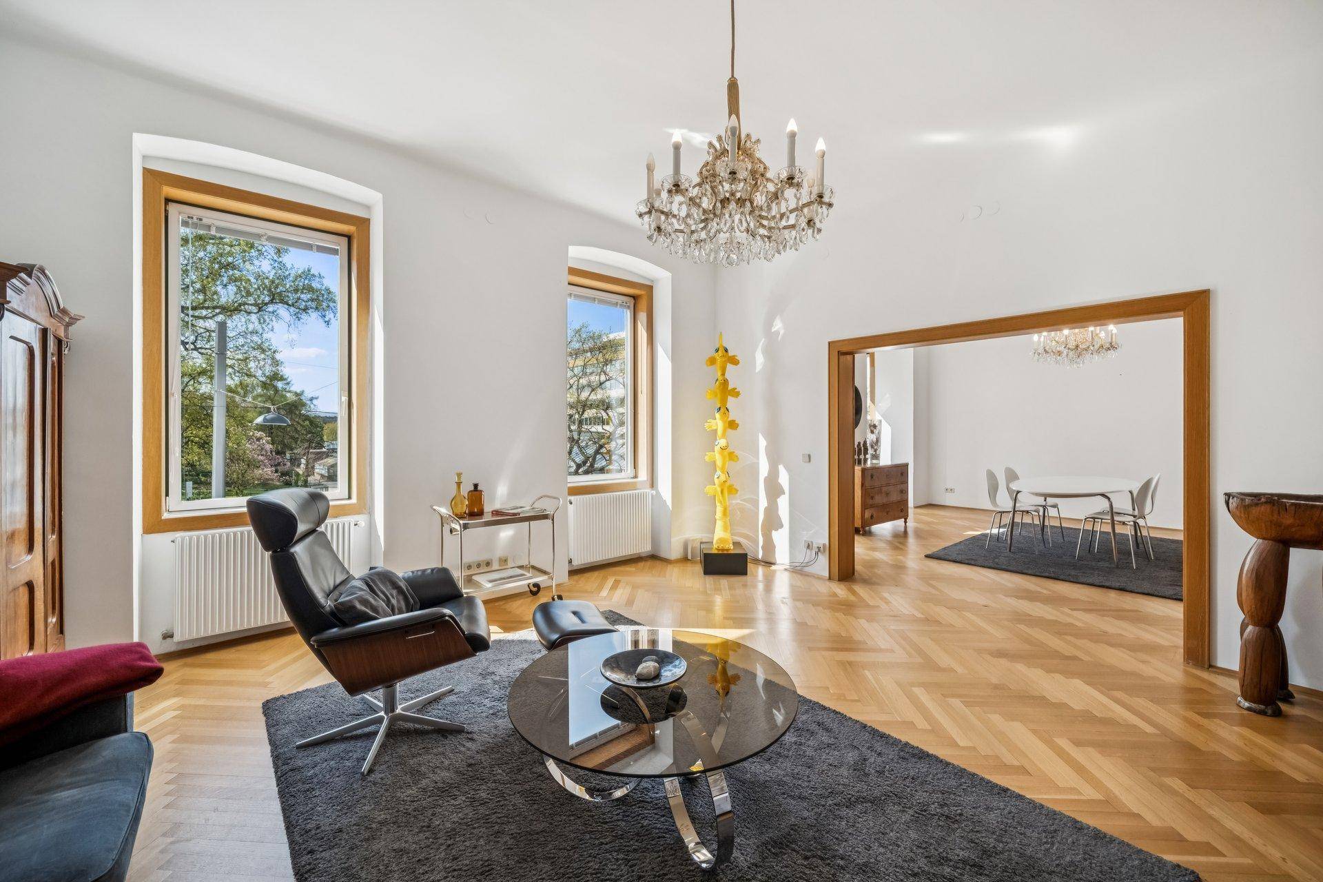 Geräumiges Wohnzimmer mit Fischgrätparkett, Kronleuchter und Blick ins Grüne durch hohe Fenster.