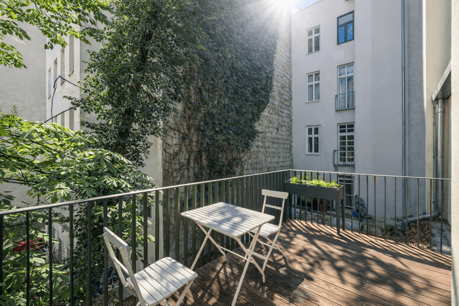 Sonniger Außenbereich mit Holzboden, Sitzgelegenheiten und Blick auf die begrünte Hausfassade.