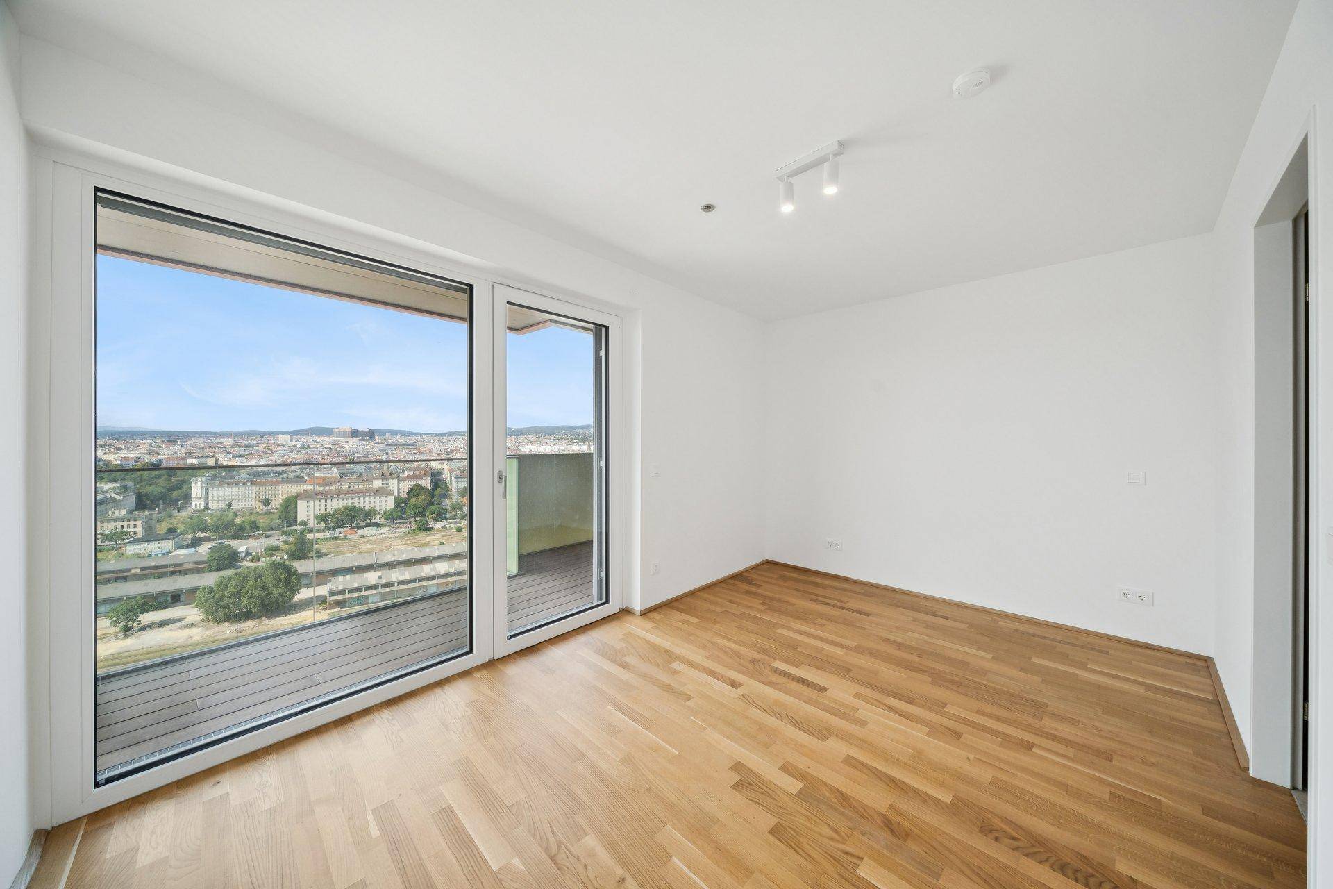 Leeres Zimmer mit Holzboden, großen Fenstern und weitem Blick über die Stadt.