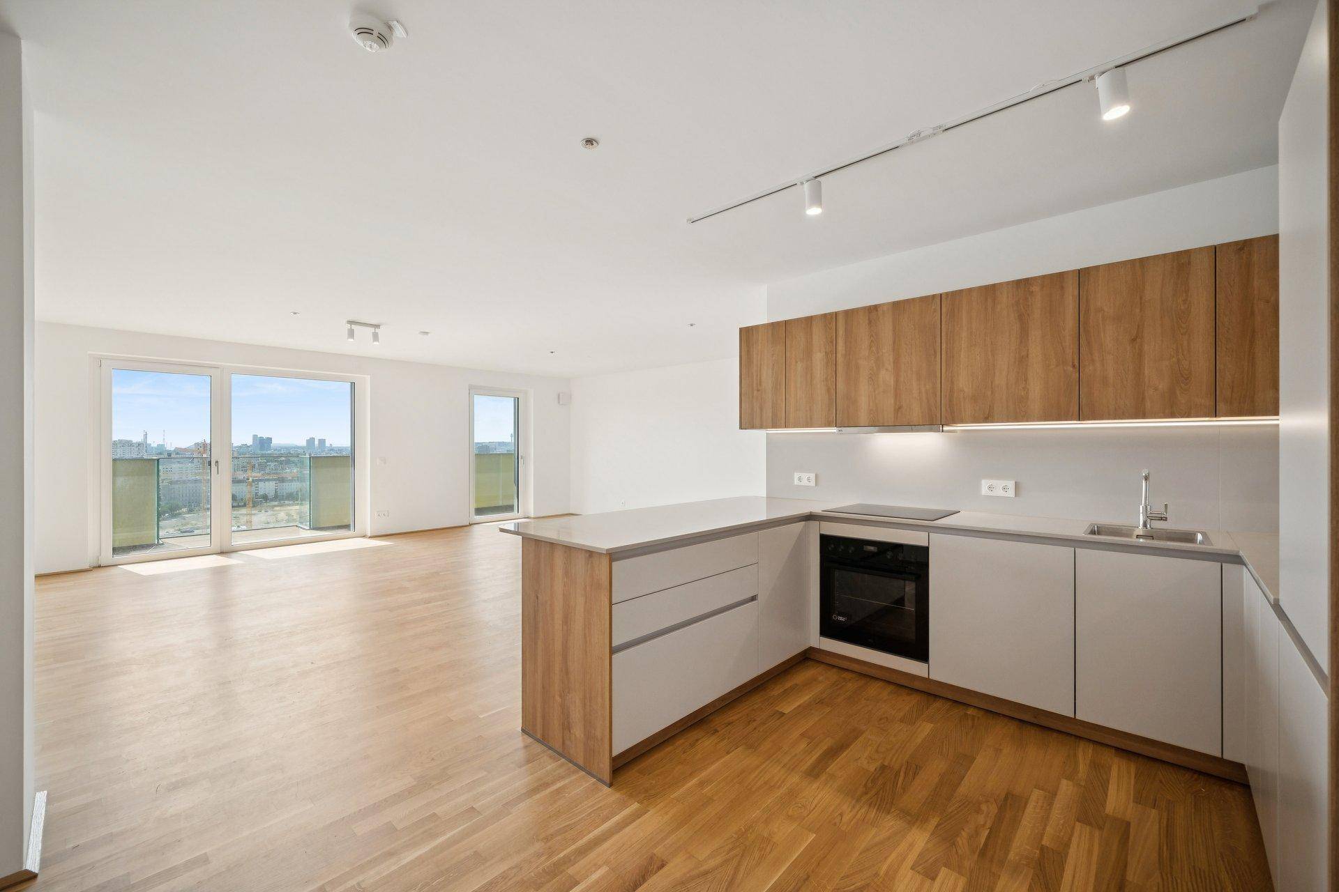 Geräumiger, leerer Wohnbereich mit offener Küche, Holzboden und Zugang zum Balkon.