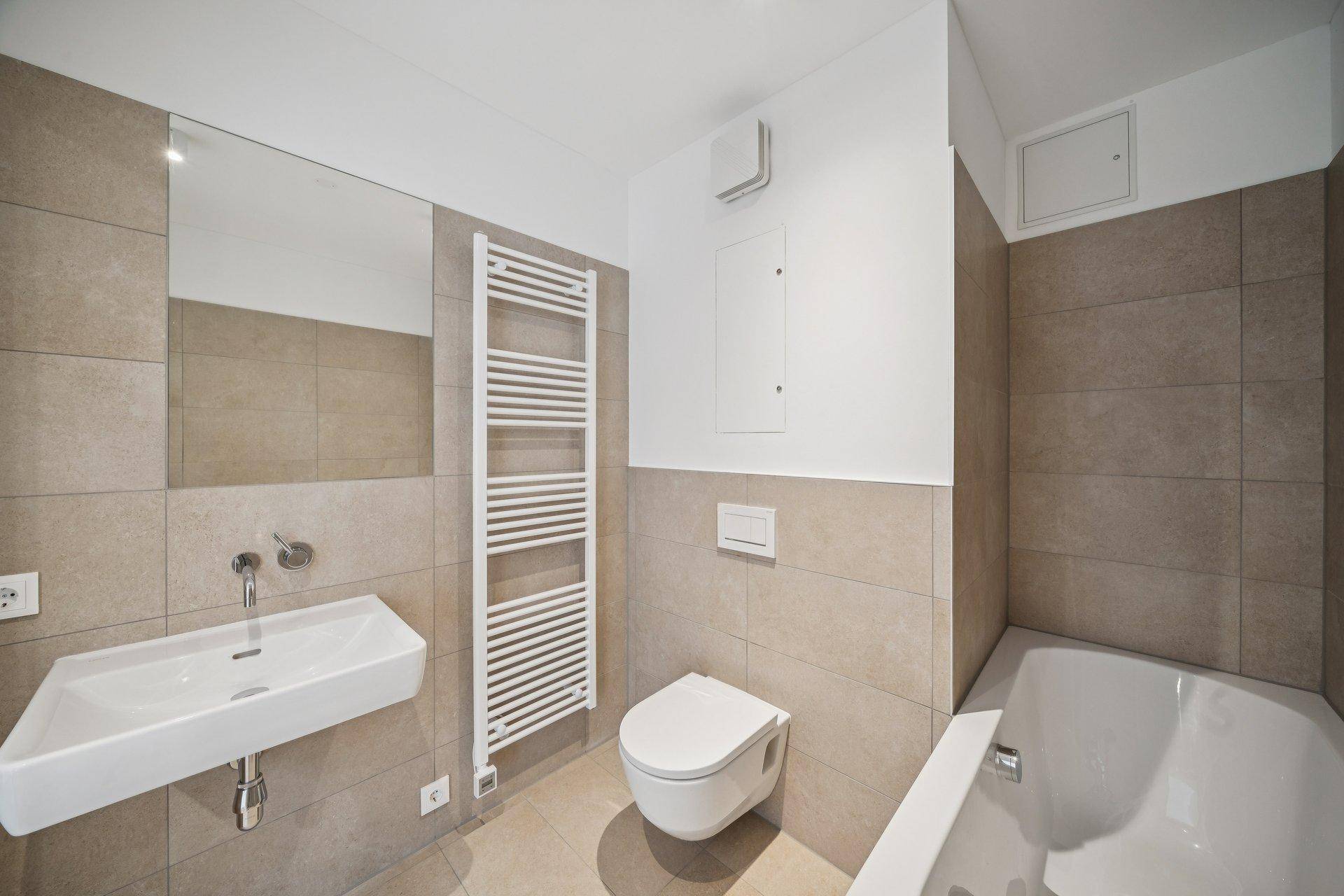 Modernes Badezimmer mit beigefarbenen Fliesen, Badewanne, Wand-WC, Einzelwaschbecken und Handtuchheizkörper.
