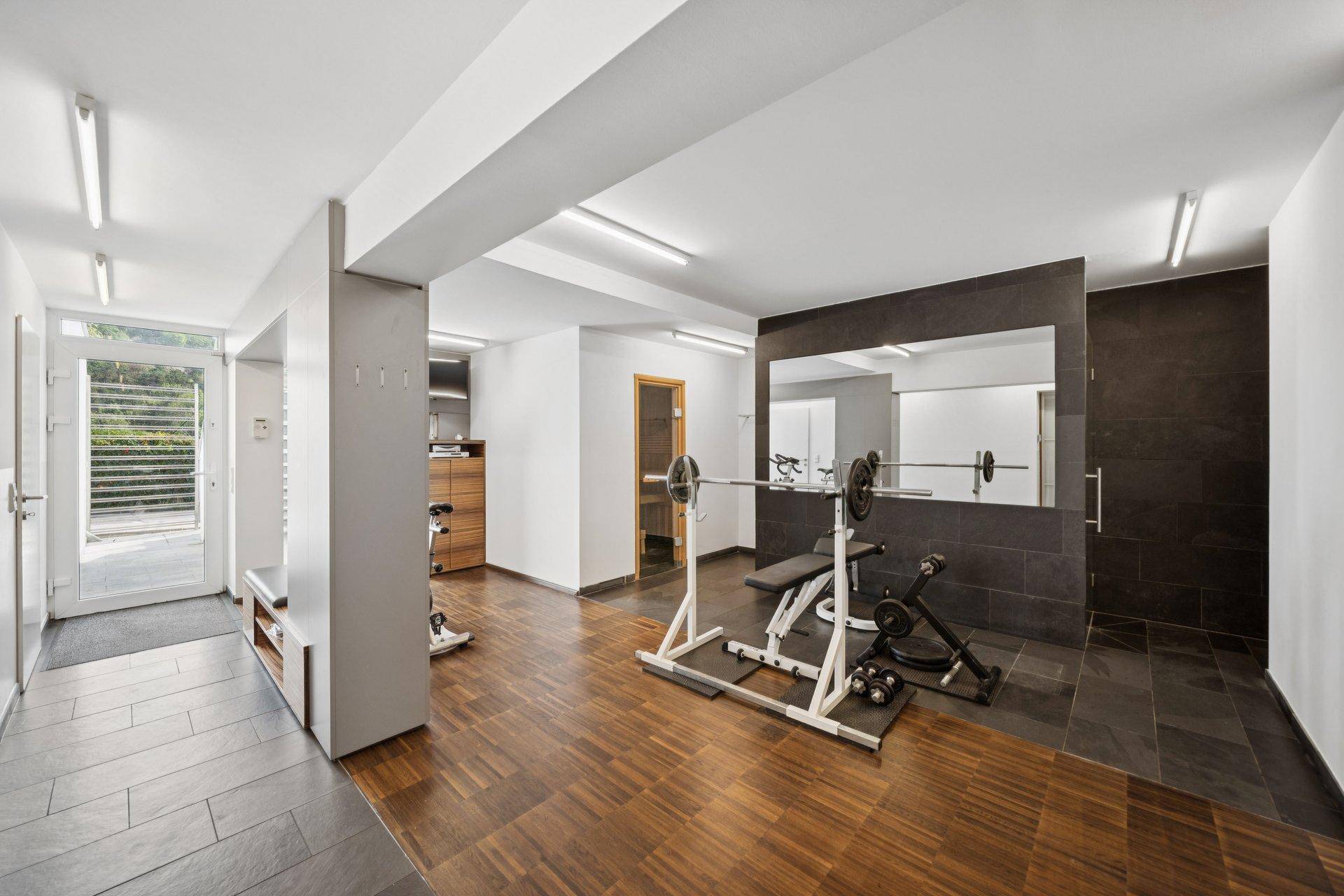 Geräumiger Fitnessraum mit verschiedenen Trainingsgeräten und Zugang zum Außenbereich.