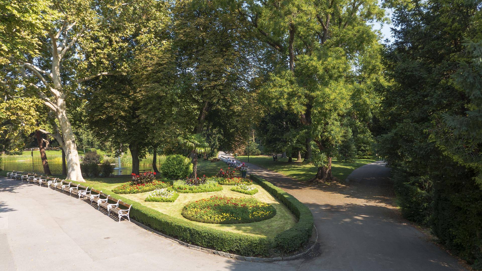 Malerischer Park mit alten Bäumen, Blumenbeeten und Sitzbänken an einem sonnigen Tag.