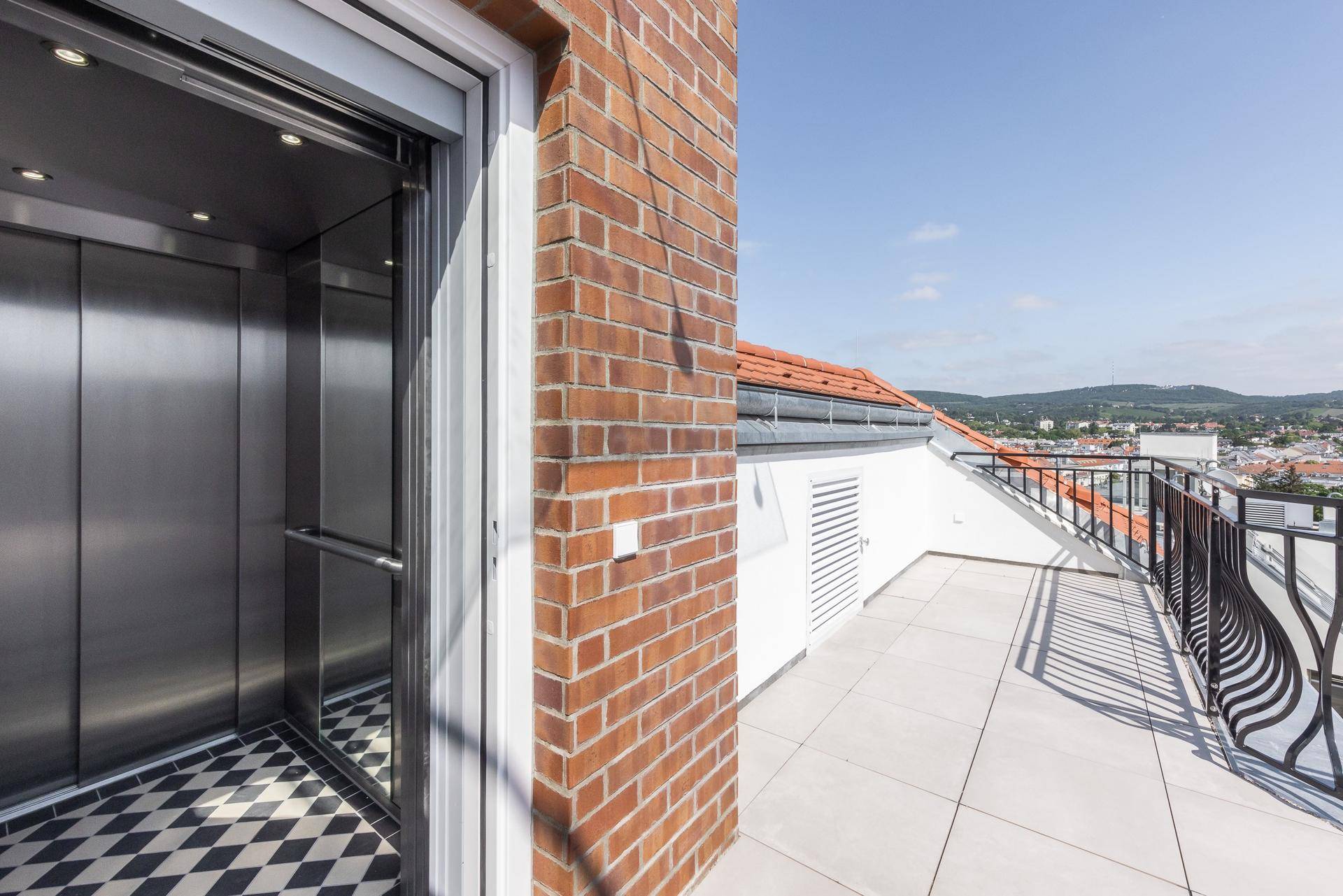 Moderner Aufzug öffnet sich zu einer geräumigen Dachterrasse mit Ziegelwand und Stadtblick.