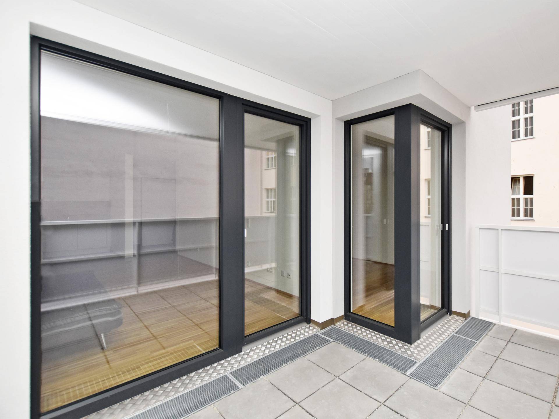 Heller Balkon mit großen Glasschiebetüren und grauem Fliesenboden, der Zugang zum Innenbereich bietet.