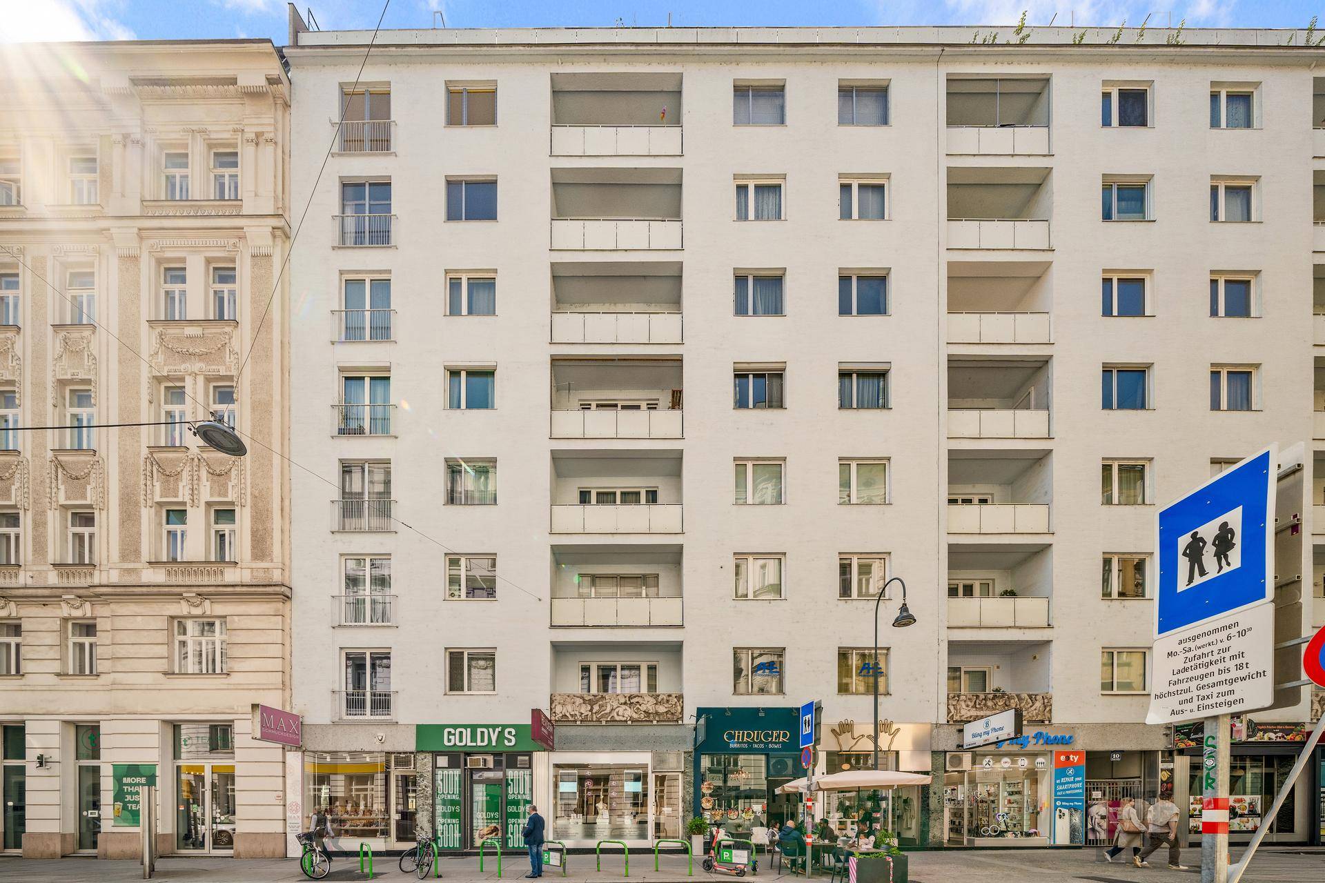 Mehrstöckiges Wohngebäude mit Balkonen und Geschäften im Erdgeschoss an einer Stadtstraße.