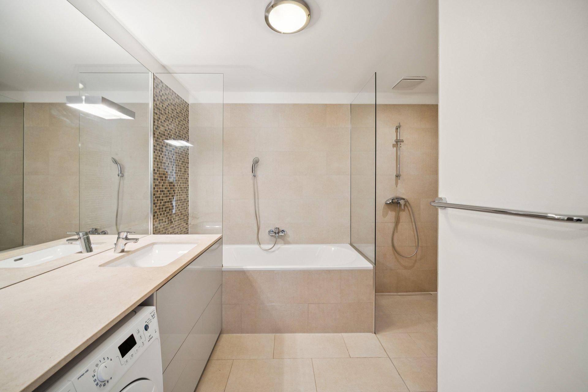 Modernes Badezimmer mit Badewanne, Dusche, beigefarbenen Fliesen und Waschmaschinenanschluss.