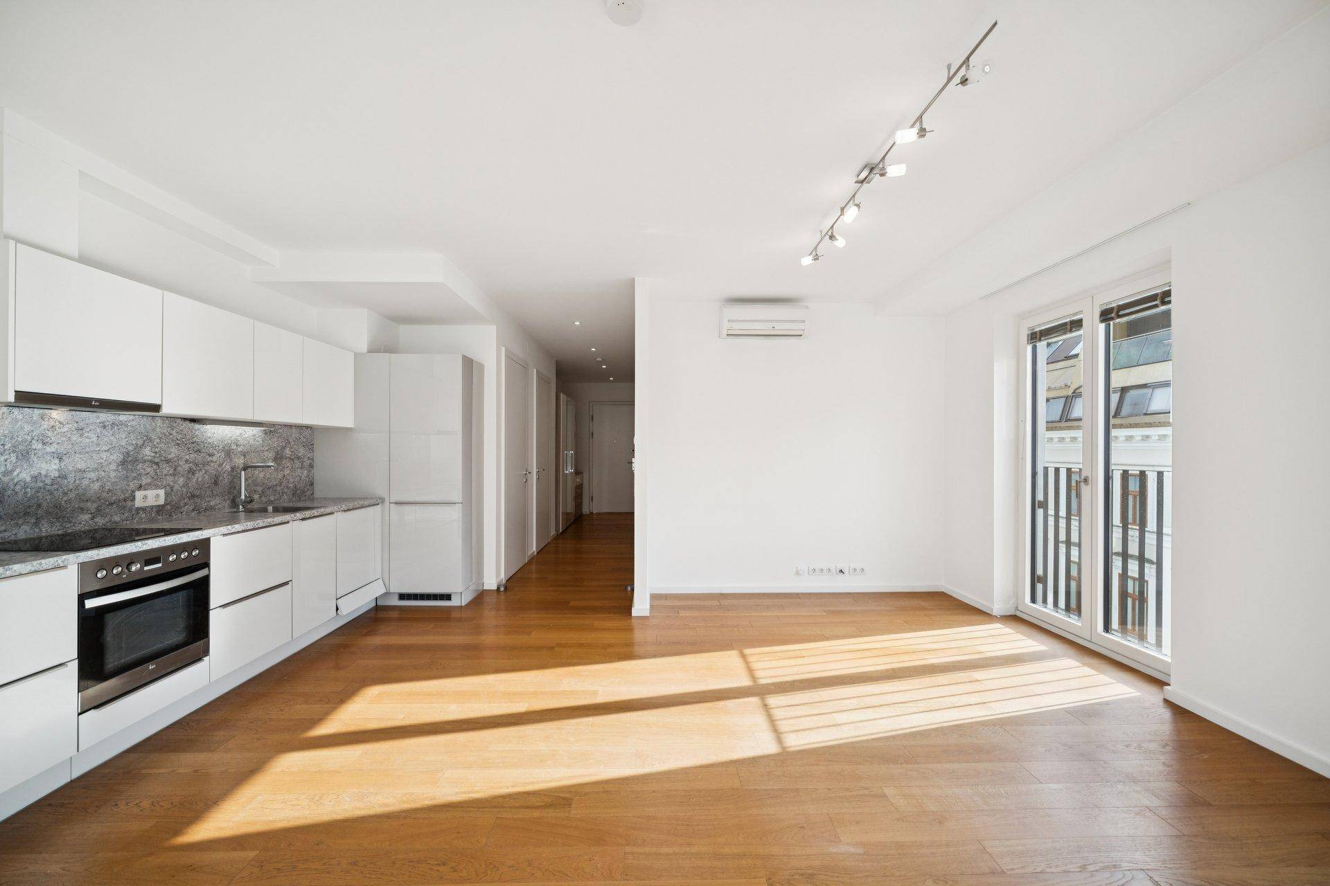 Heller, leerer Wohnbereich mit offener Küche, Holzparkett und Zugang zu einem französischen Balkon.