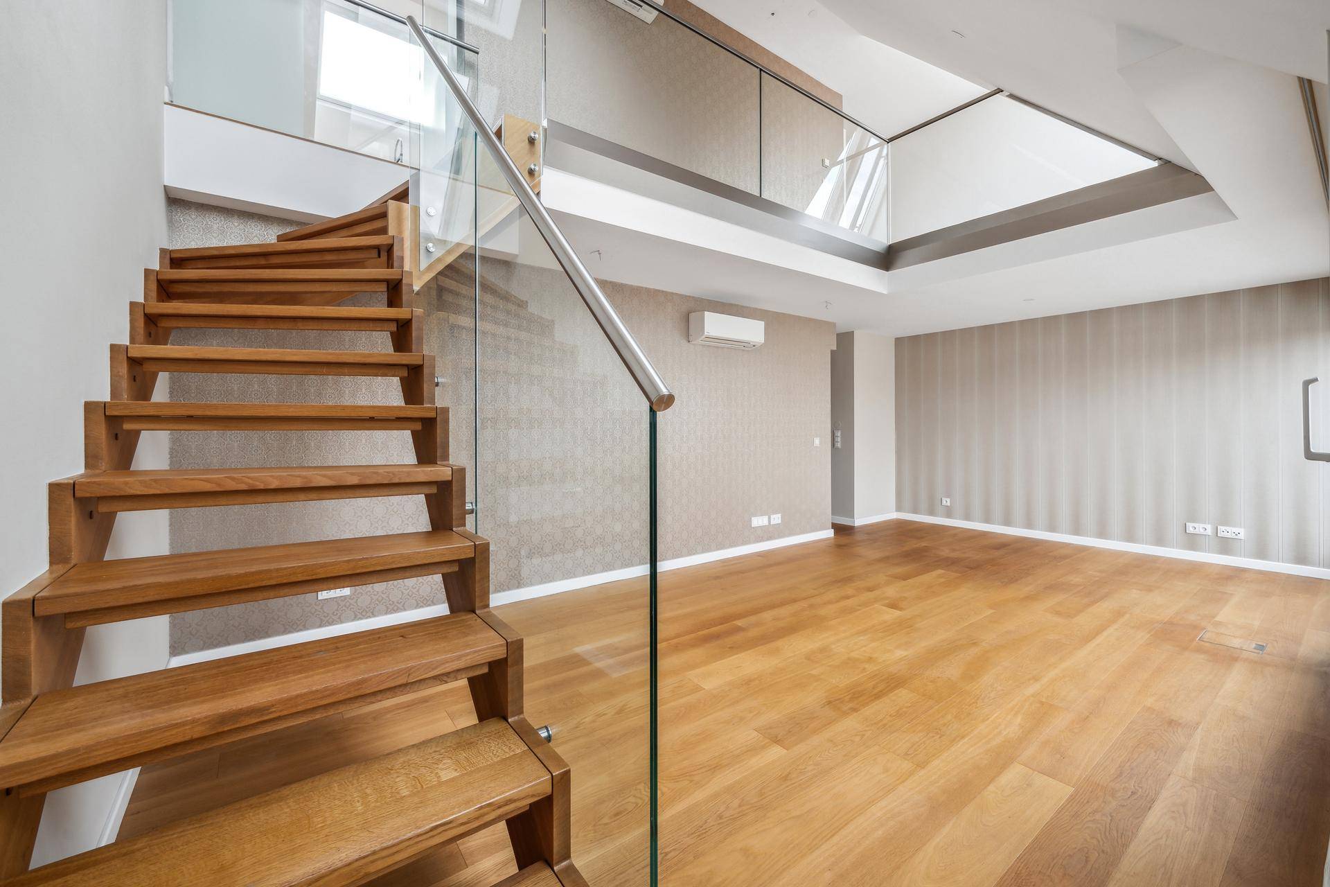 Geräumiges Wohnzimmer mit Holztreppe, Glasgeländer und Dachfenstern, bereit zur Einrichtung.