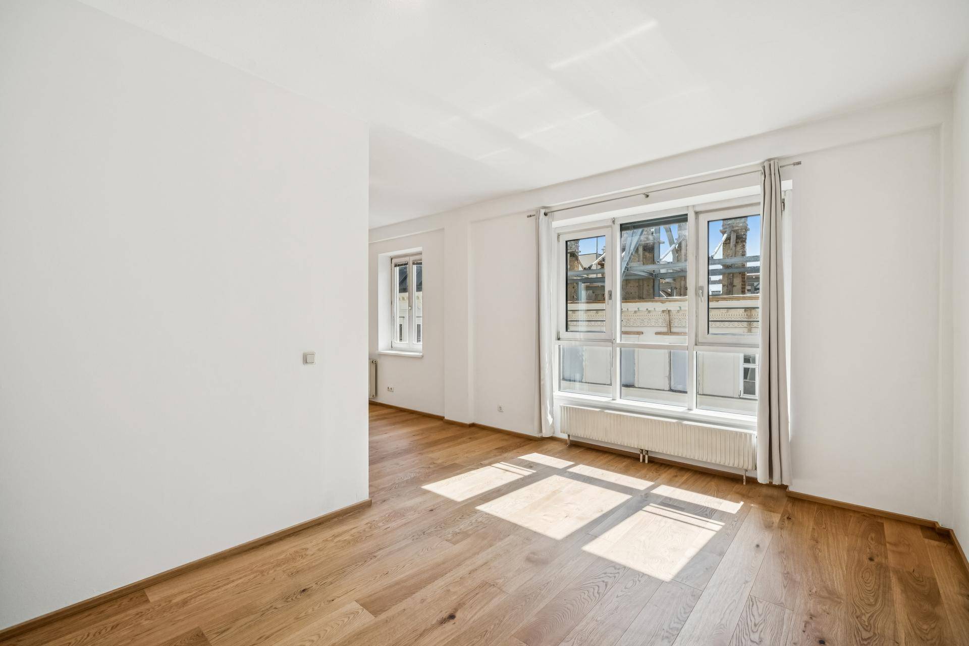 Geräumiges Wohnzimmer mit Holzboden und großen Fenstern, die den Raum hell erscheinen lassen.