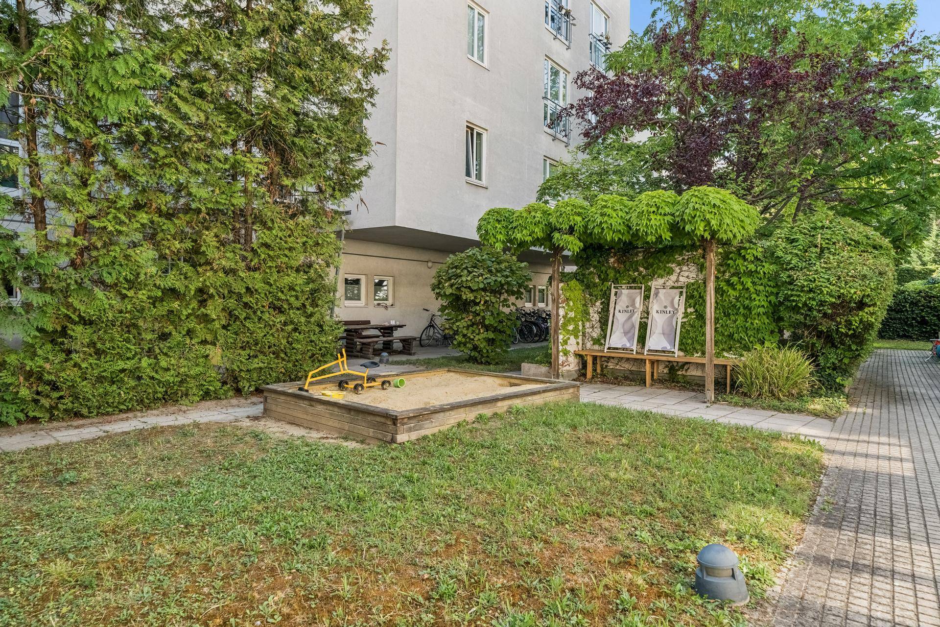 Gemeinschaftsgarten mit Sandkasten und Sitzgelegenheiten unter Bäumen für Kinder und Erwachsene.