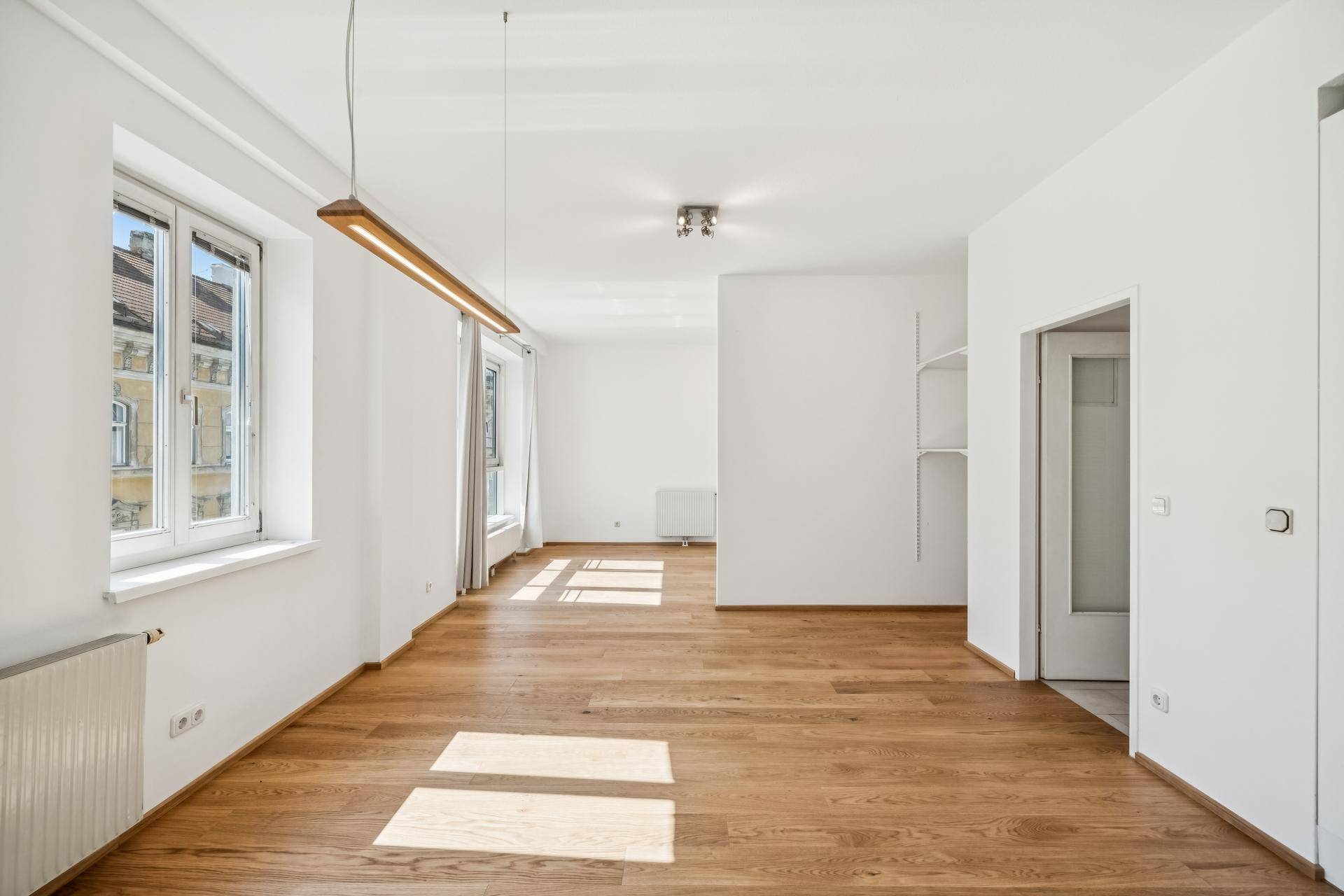 Geräumiger Wohnbereich mit Holzboden, großen Fenstern und integrierten Regalen.