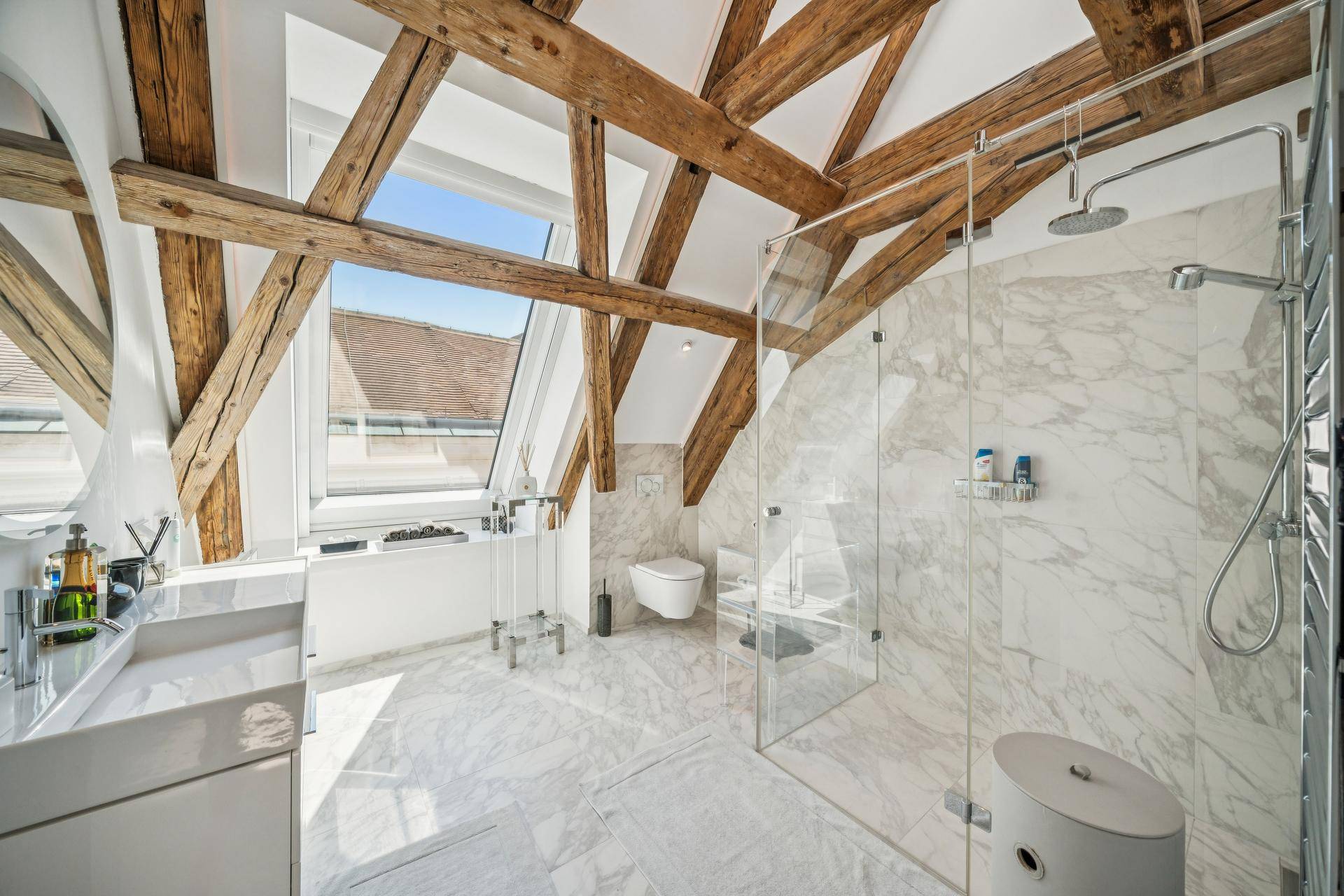 Modernes Badezimmer mit Marmorfliesen, begehbarer Dusche und freiliegenden Holzbalken unter einem Dachfenster.