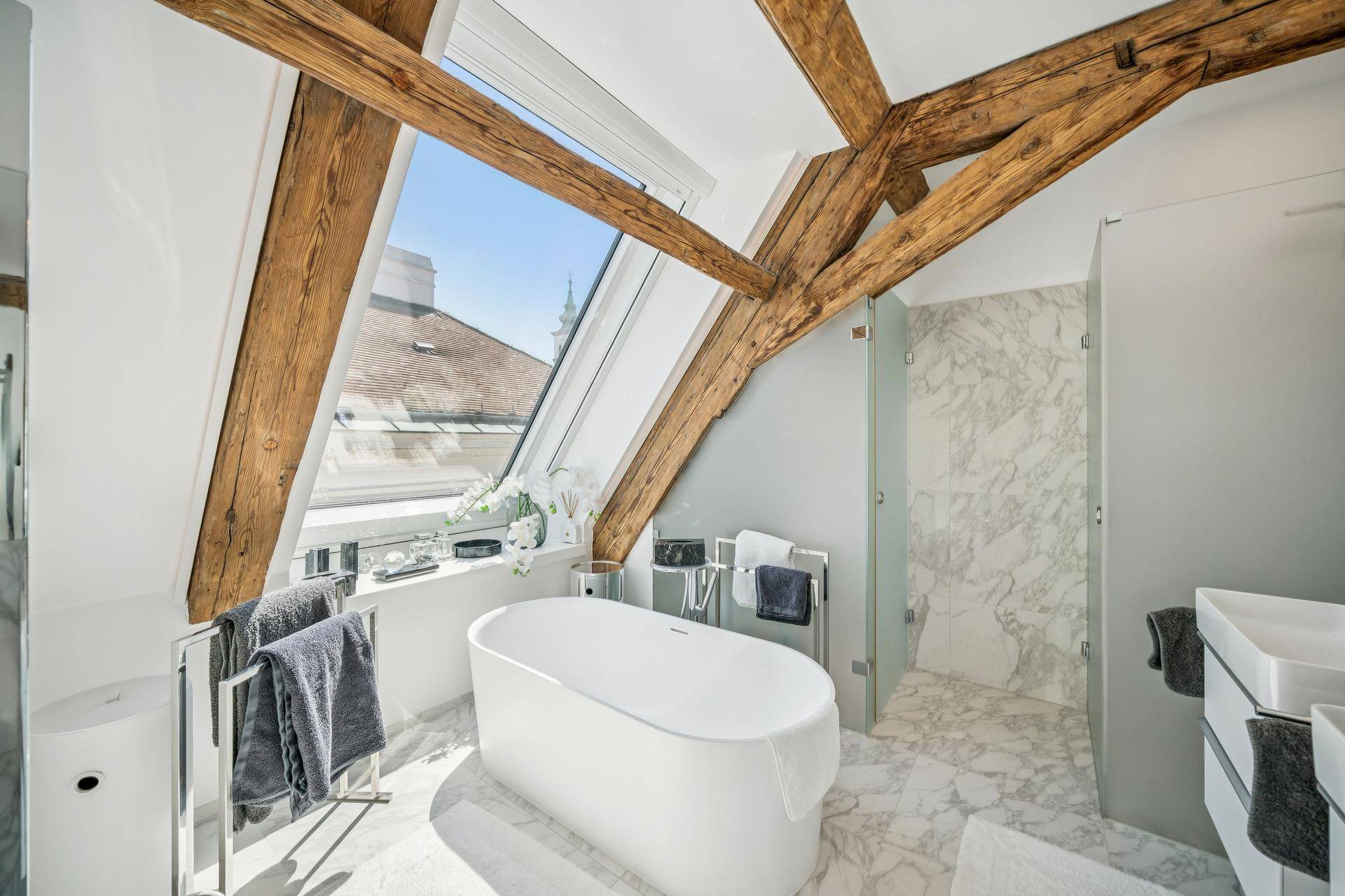Luxuriöses Badezimmer mit freistehender Badewanne, Marmorfliesen und Doppelwaschbecken unter einem Dachfenster.