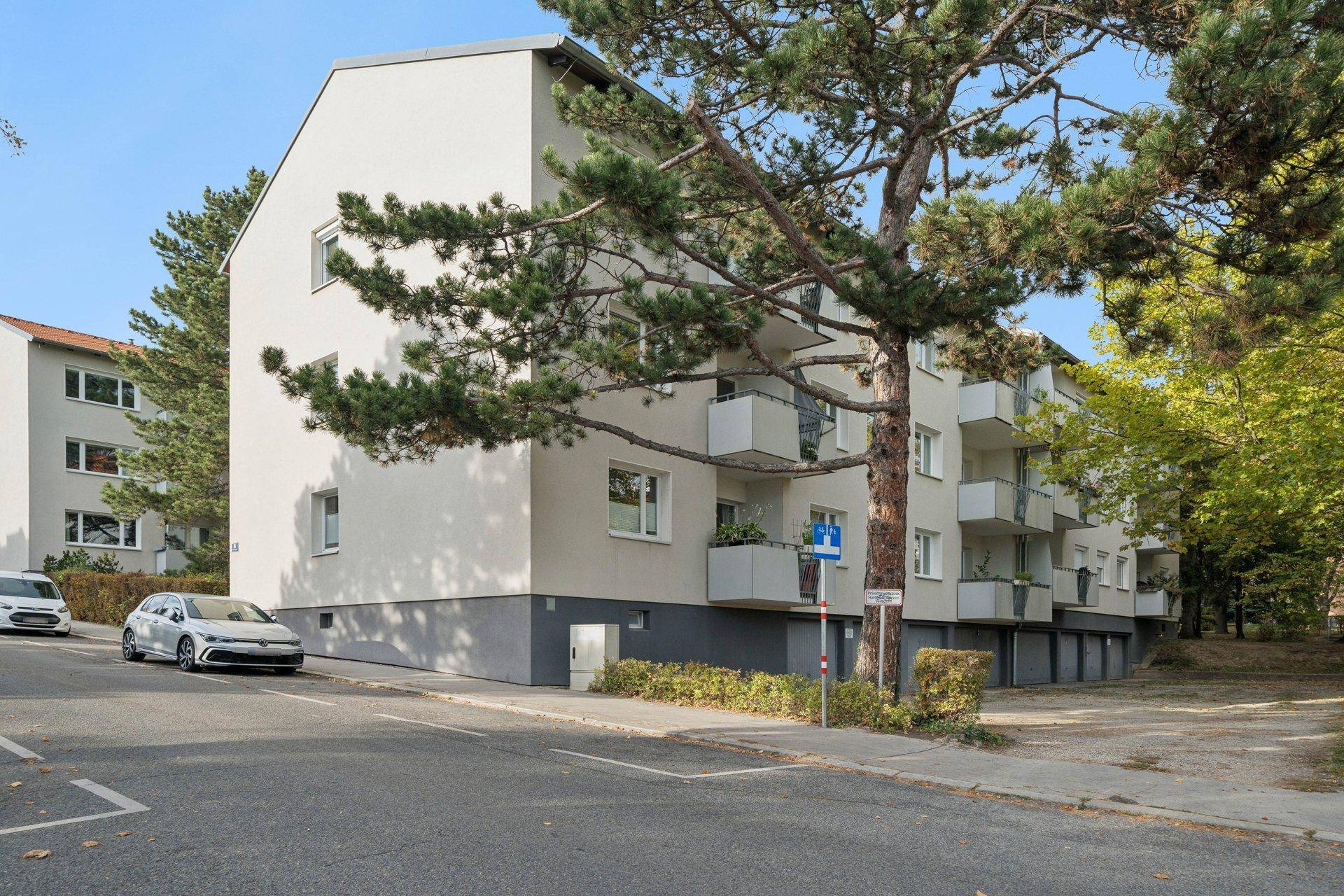Mehrfamilienhaus mit moderner Fassade, Balkonen und umgebenden Bäumen an einer Straße.