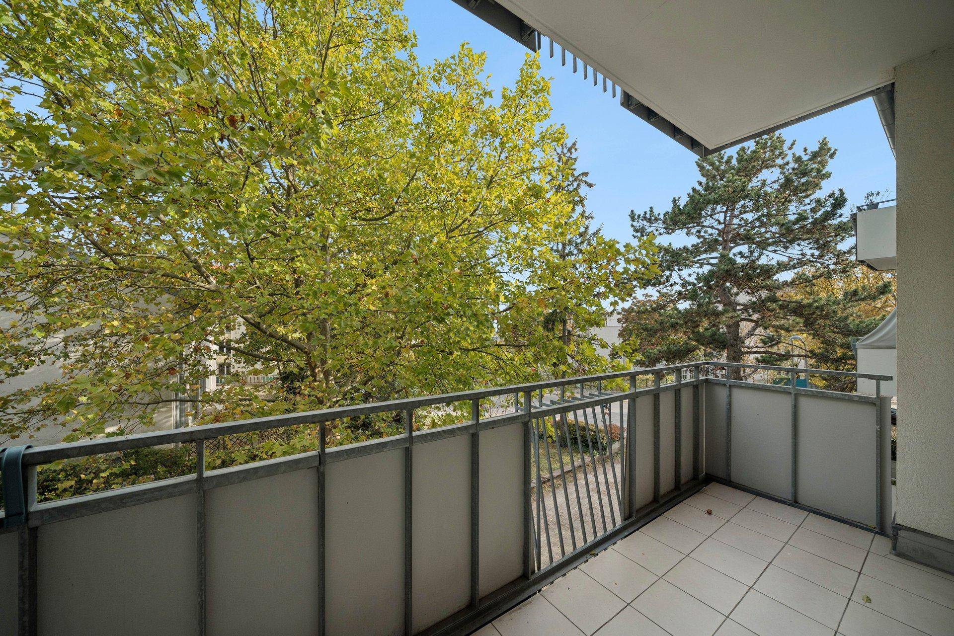 Leerer Balkon mit Fliesenboden und Metallgeländer, umgeben von Bäumen.