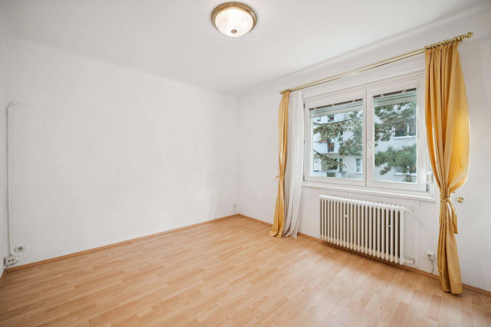 Helles Schlafzimmer mit Parkettboden, Heizkörper und gelben Vorhängen am Fenster.