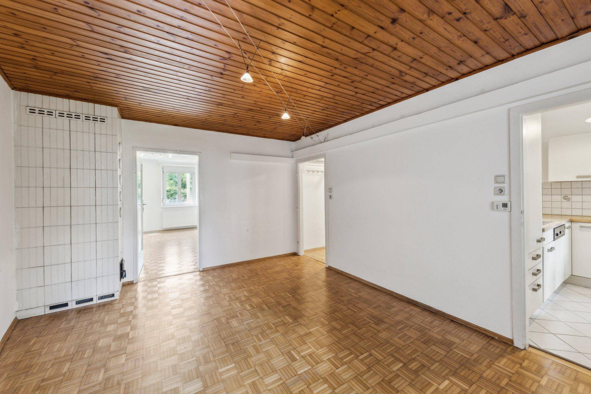 Geräumiges Zimmer mit Holzbalkendecke, Kachelofen und Zugang zu weiteren Räumen.