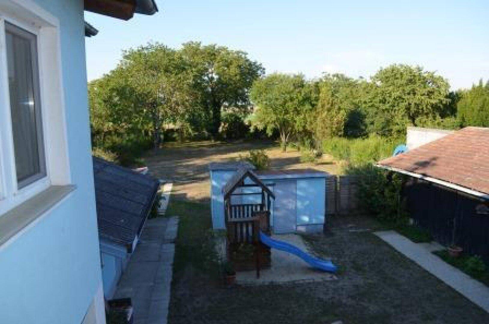 Blick aus dem Fenster auf den Garten mit Spielplatz und weiteren Bäumen im Hintergrund.