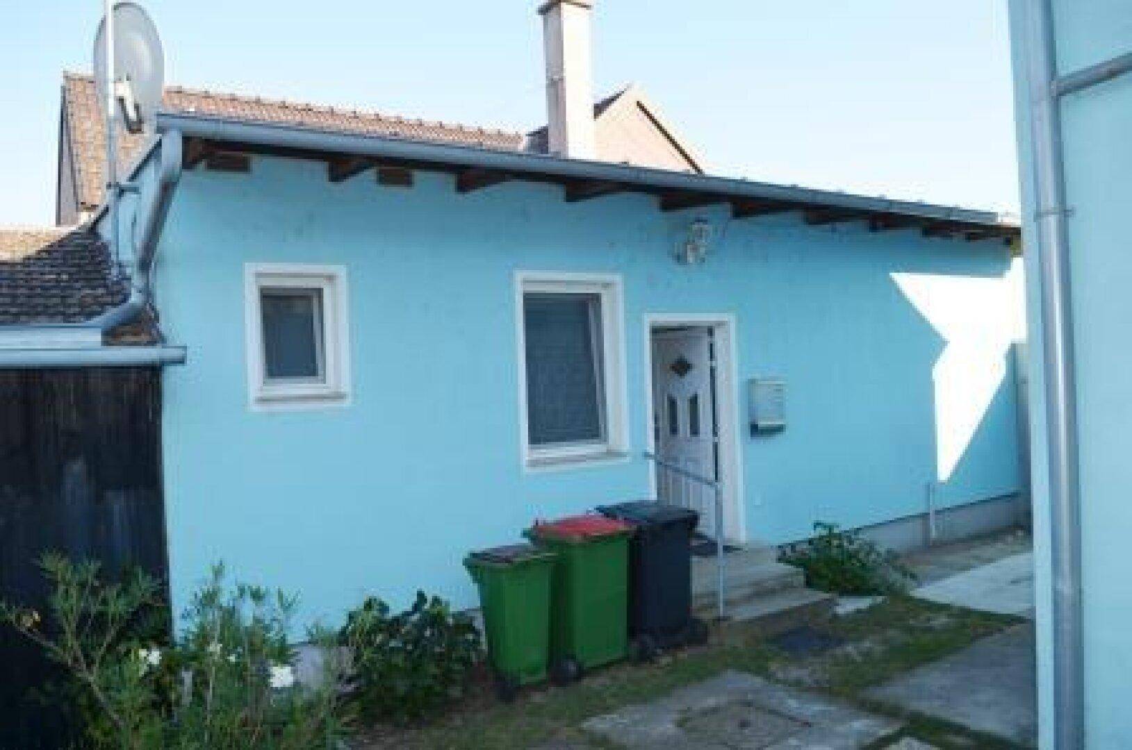 Eingangsbereich des blauen Hauses mit Mülltonnen und gepflastertem Zugang.