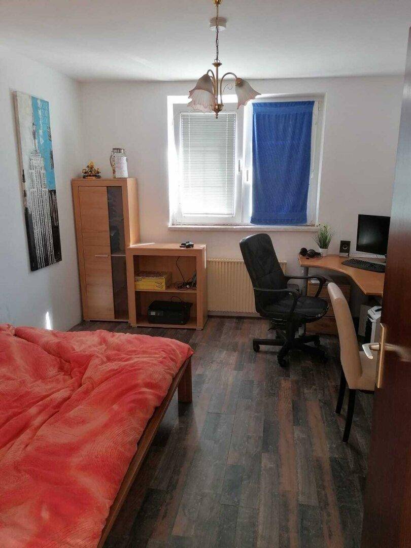 Gemütliches Schlafzimmer mit Holzbett, Schreibtisch und dunklem Laminatboden, ideal für Wohnen und Arbeiten.