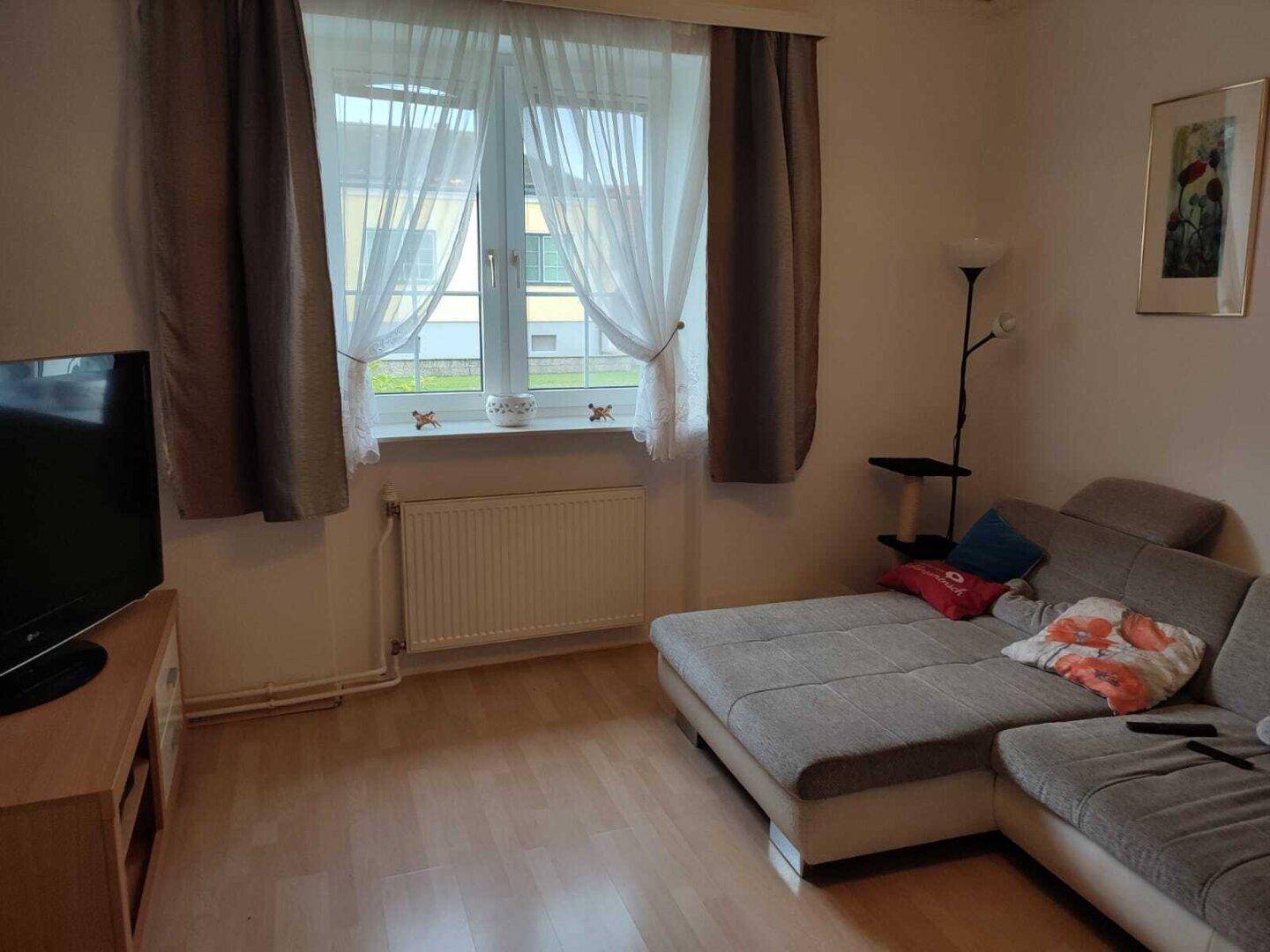 Wohnzimmer mit beigem Ecksofa, großem Fenster und Heizkörper unter dem Fenster.