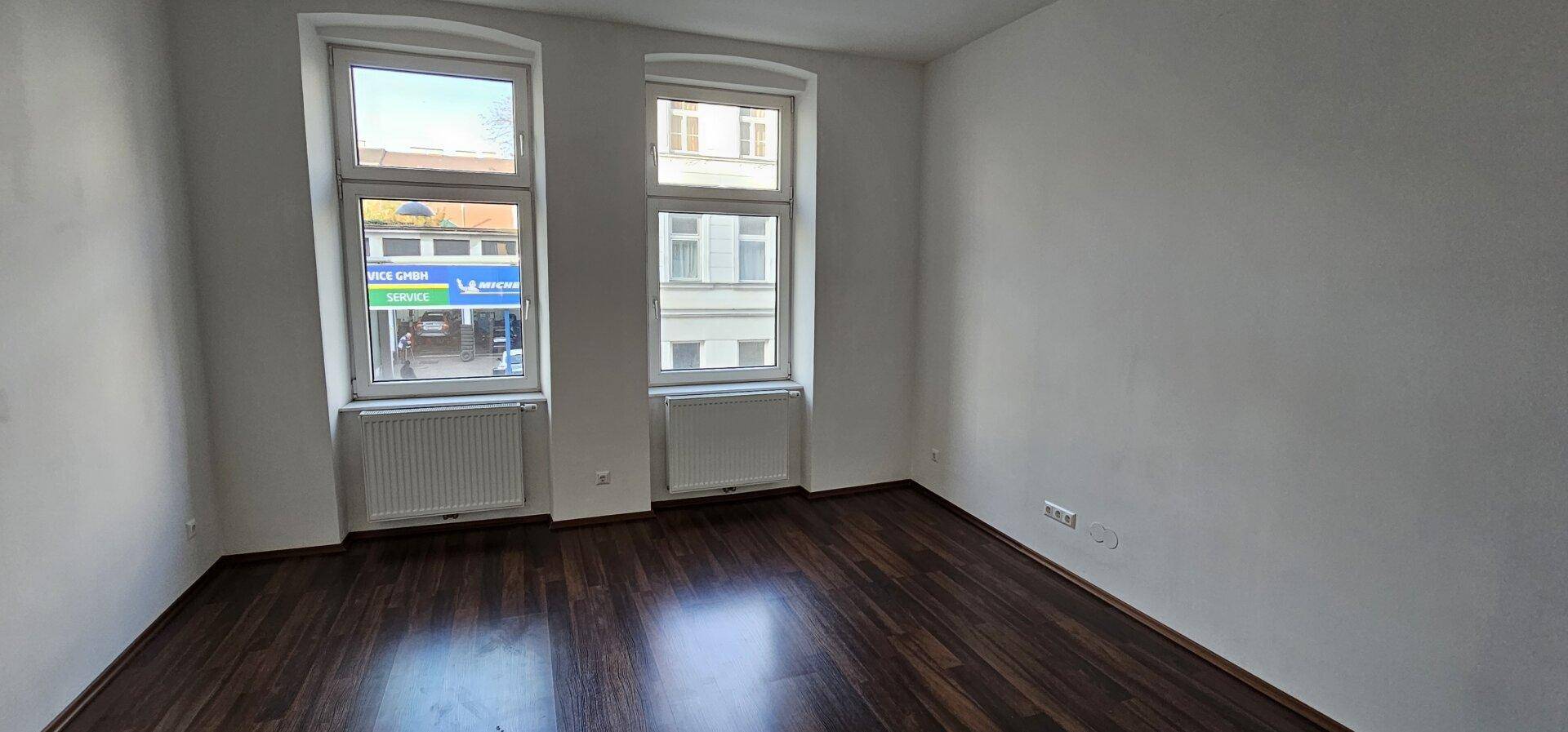 Leerer Raum mit dunklem Holzboden, weißen Wänden und zwei hohen Fenstern.
