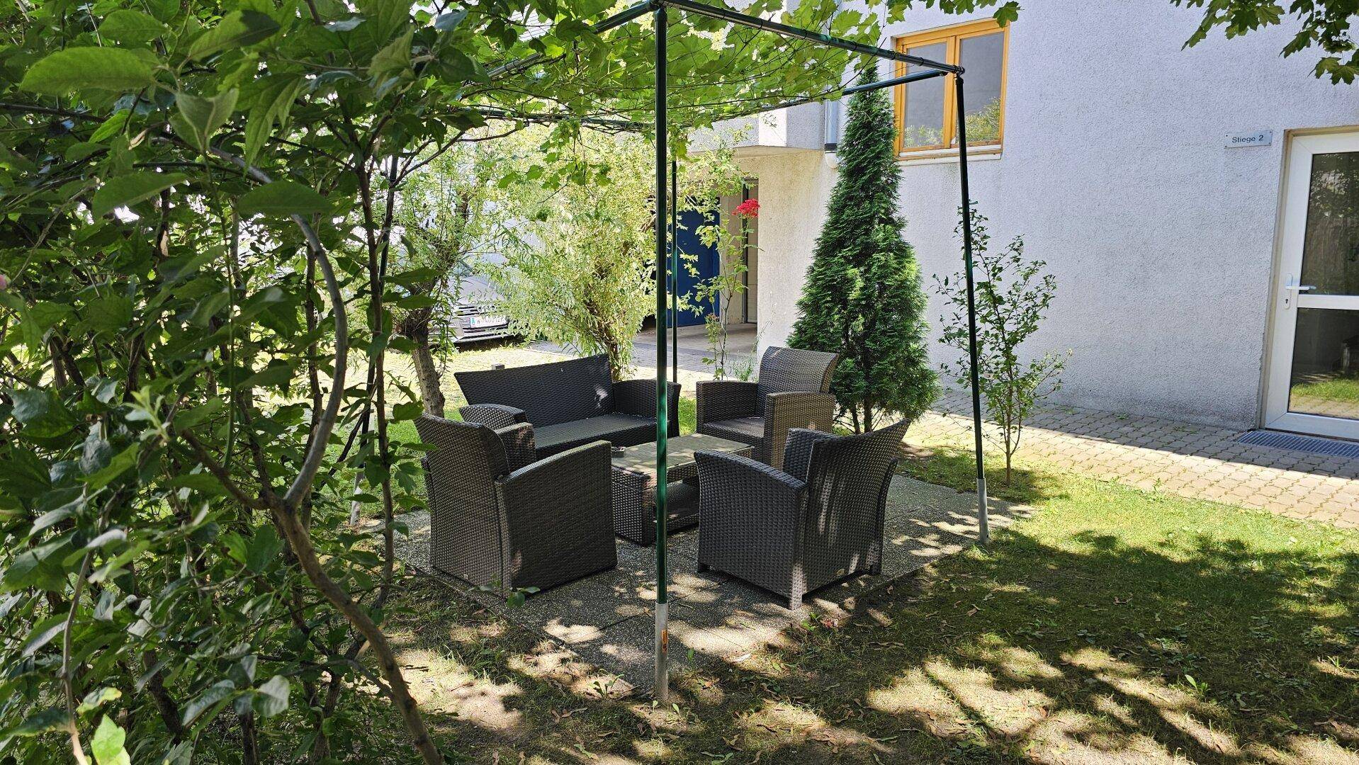 Gemütlicher Sitzbereich im Garten unter einem grünen Gestell, umgeben von Bäumen.