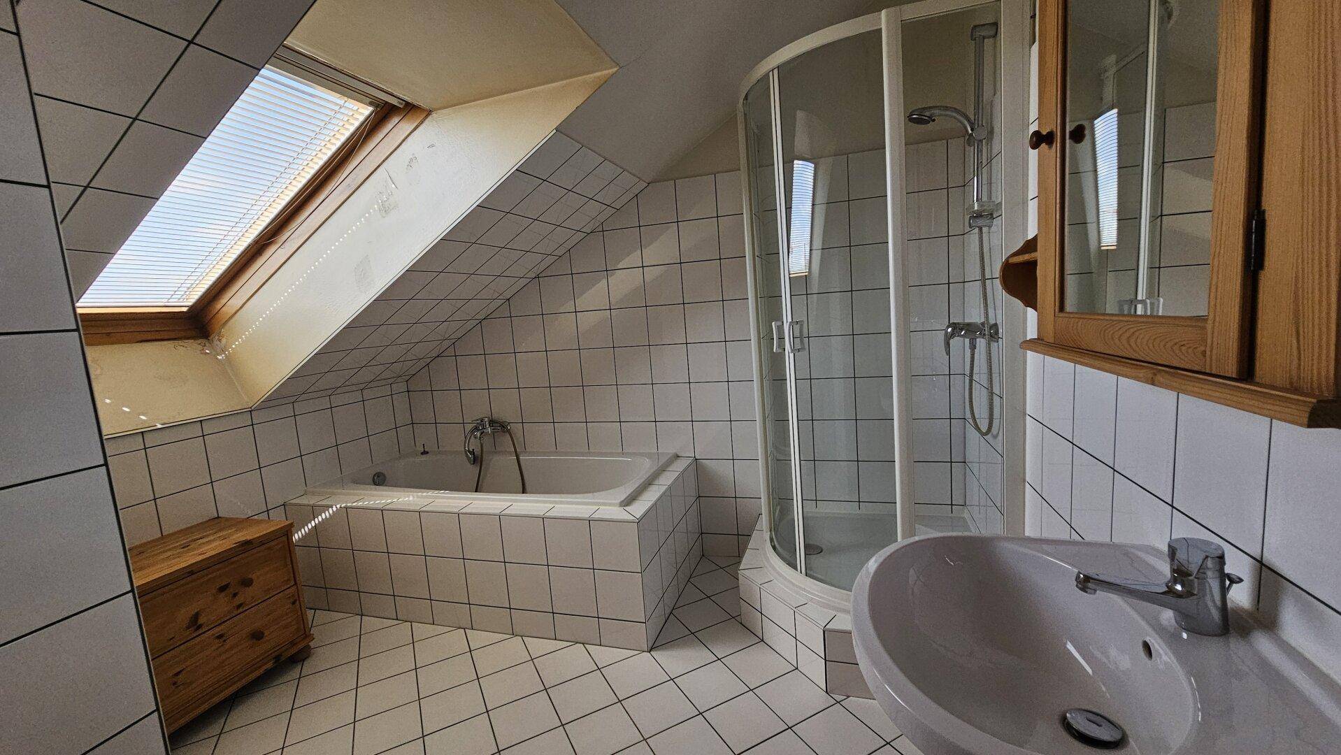 Helles Badezimmer mit weißen Fliesen, Duschkabine und Badewanne unter dem Dachfenster.