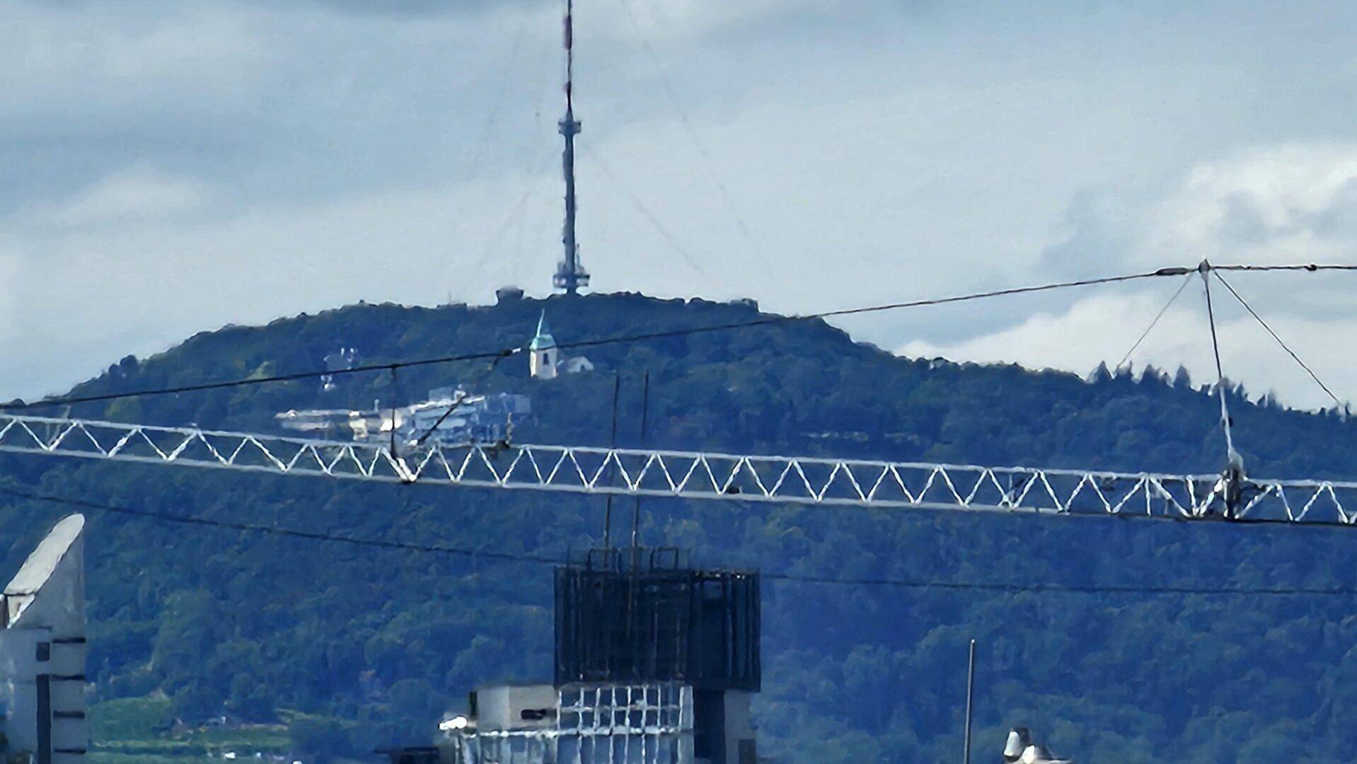 Weitblick auf einen bewaldeten Berg mit Sendeturm und Gebäuden unter bewölktem Himmel.