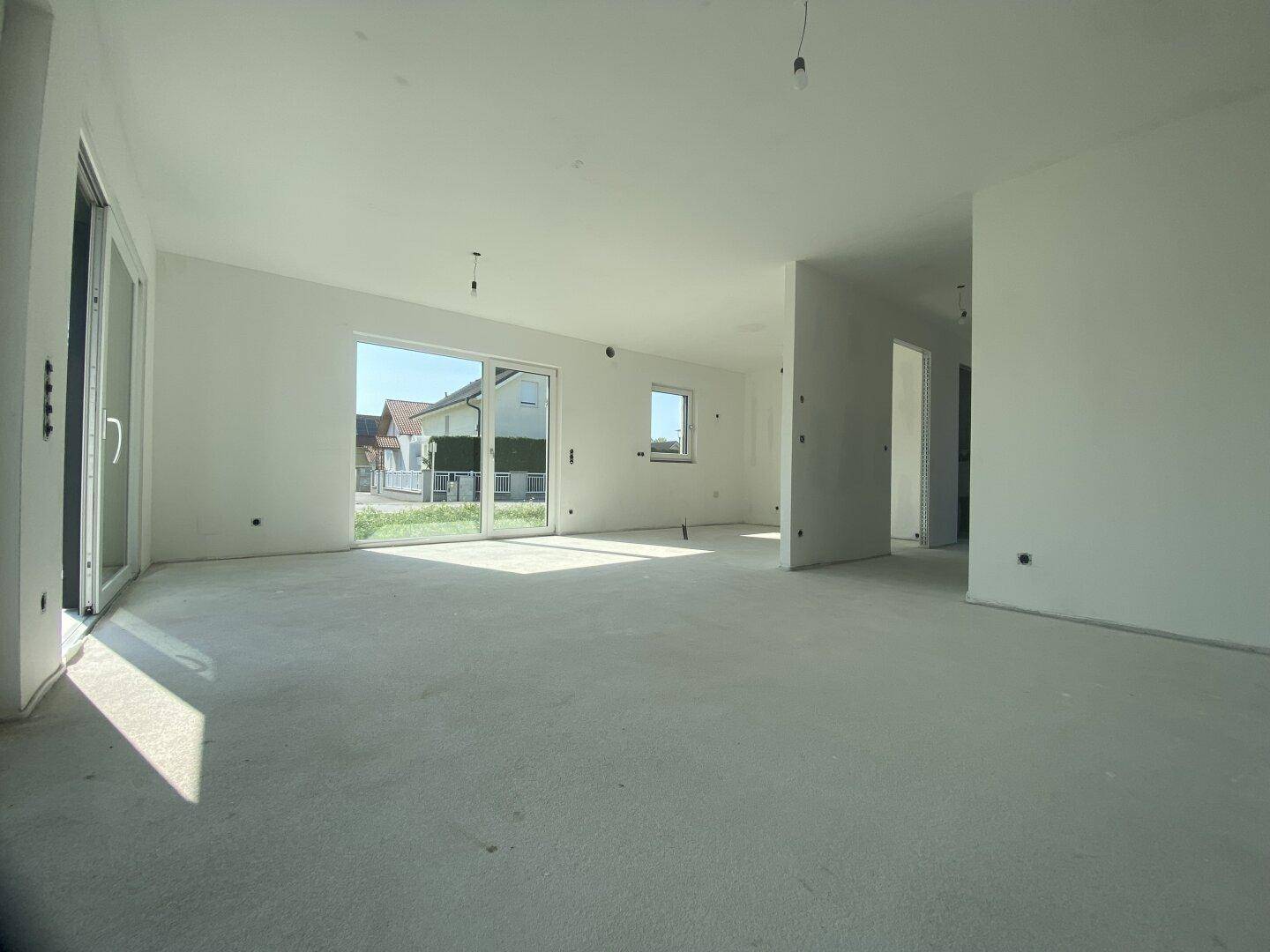 Geräumiger, unfertiger Wohnbereich mit großen Fenstern und Betonboden, bereit für den Ausbau.