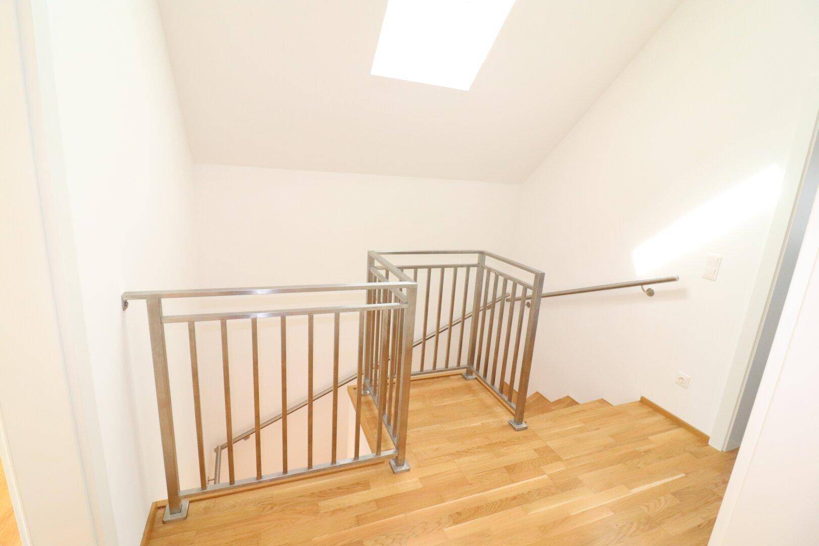 Heller Treppenaufgang mit Holzstufen, Edelstahlgeländer und Dachfenster.