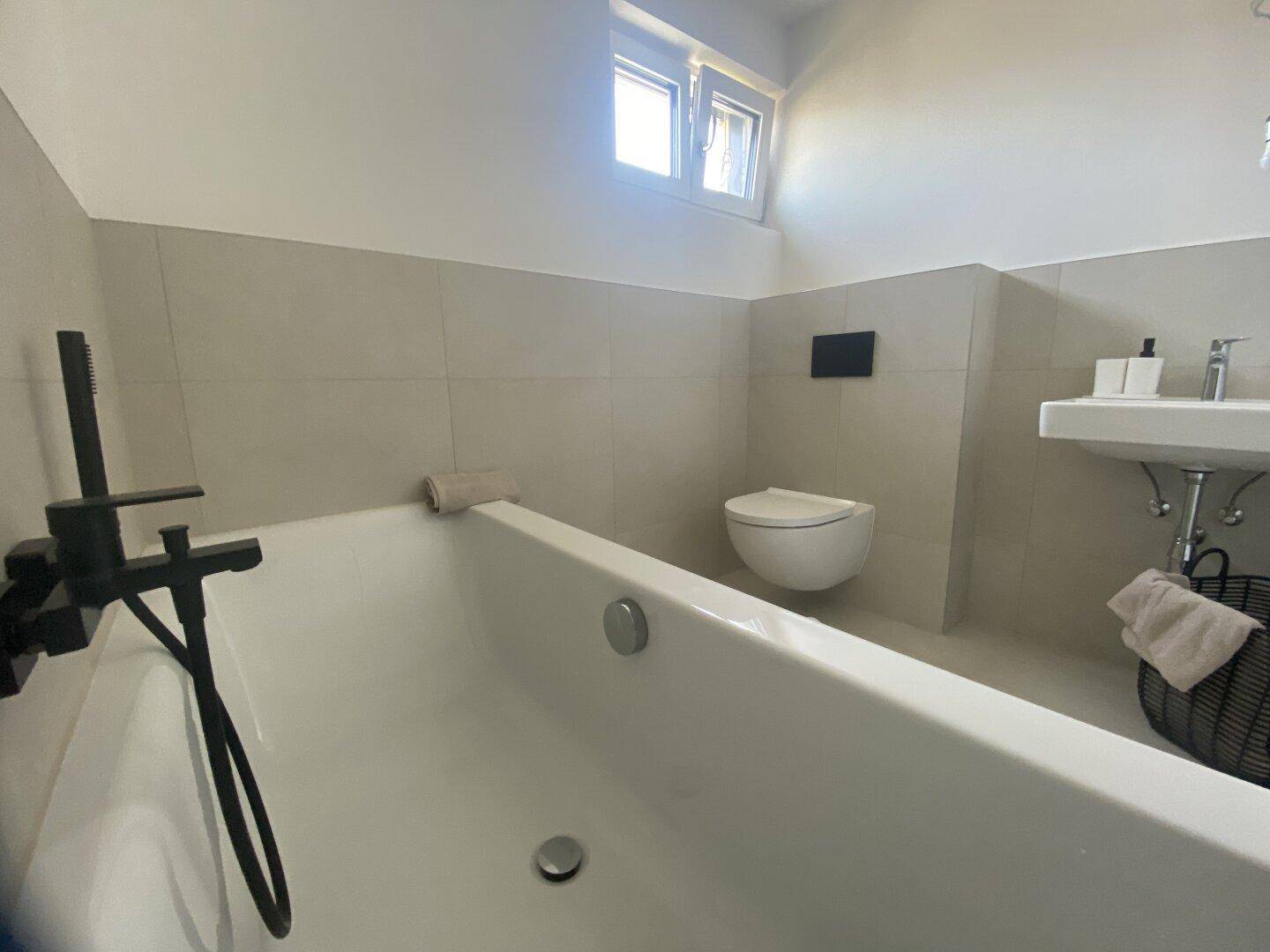 Modernes Badezimmer mit Badewanne, Wand-WC, Einzelwaschbecken und einem Fenster für Tageslicht.