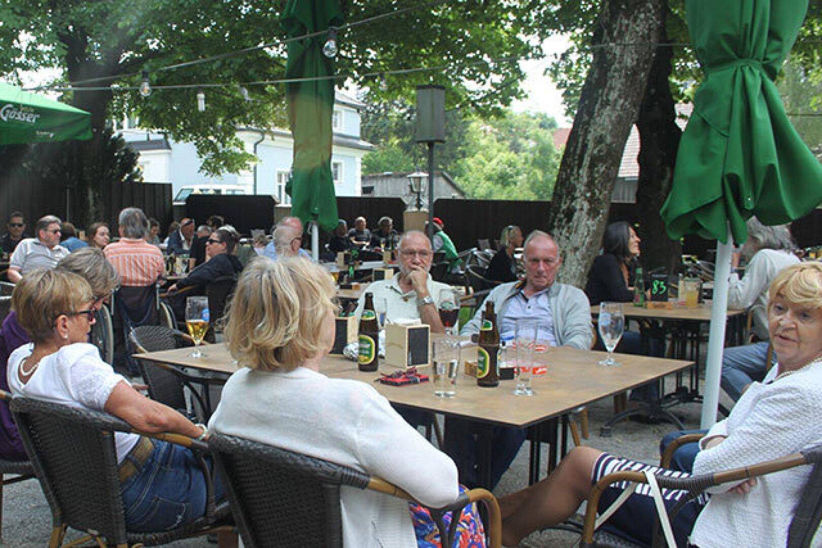 Gäste entspannen sich in einem Biergarten unter Bäumen und grünen Sonnenschirmen an einem sonnigen Tag.