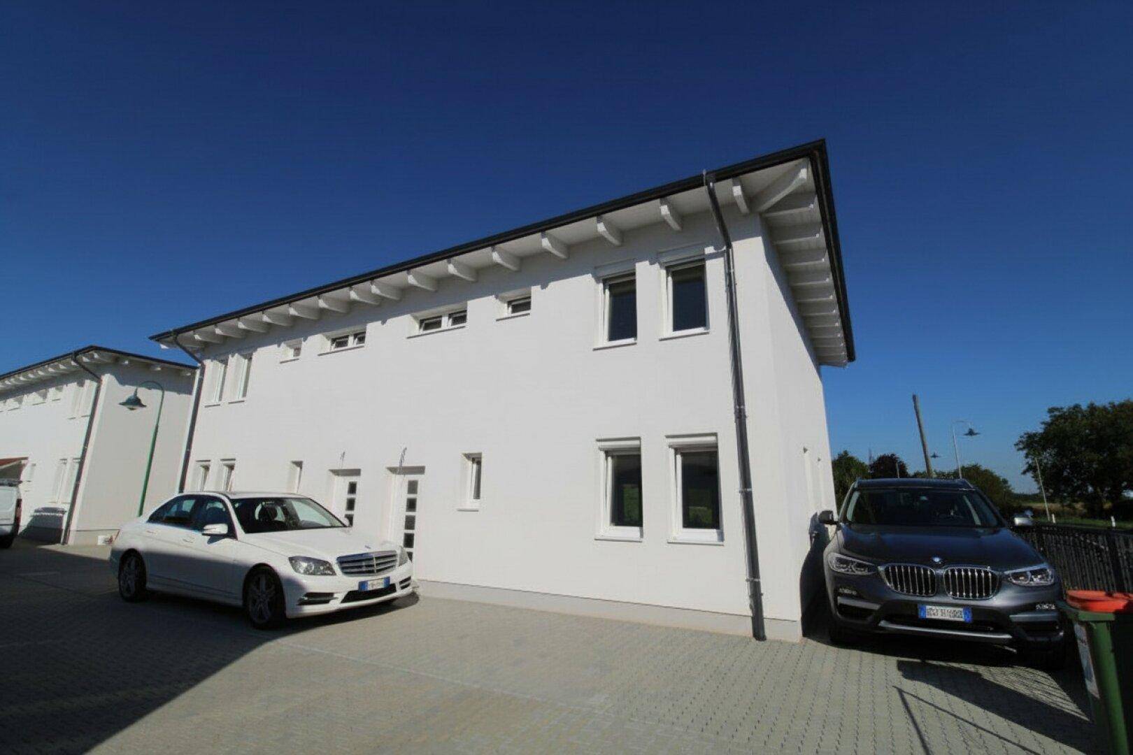 Moderne weiße Hausfassade mit klaren Linien und gepflasterter Auffahrt, zwei Autos parken davor.
