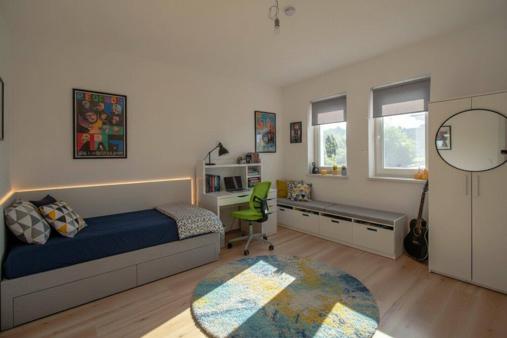 Jugendzimmer mit Bett, Schreibtisch, Stauraum und farbenfrohem Teppich, hell und funktional eingerichtet.