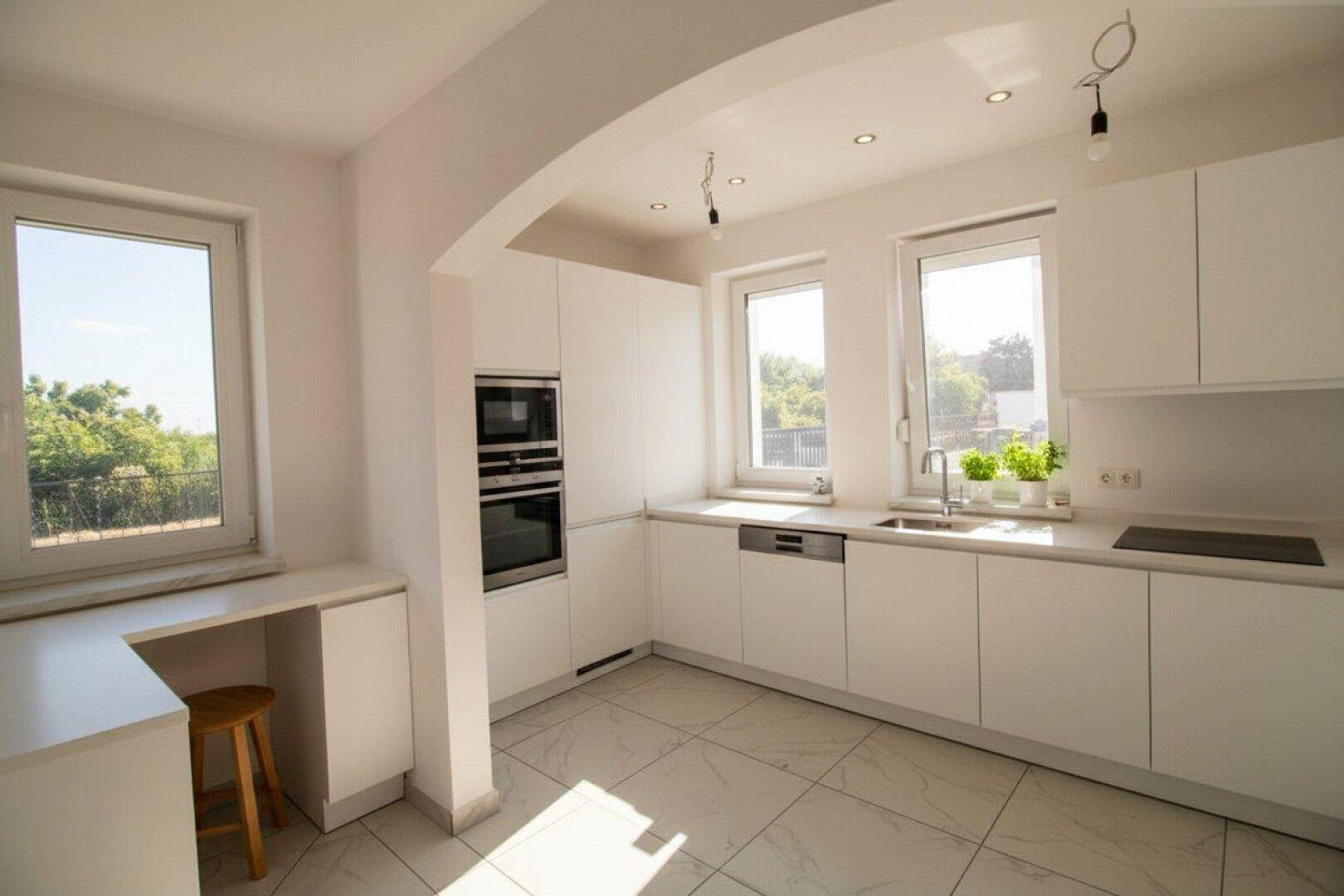 Moderne weiße Einbauküche mit integrierten Geräten, Fliesenboden und viel Tageslicht durch Fenster.