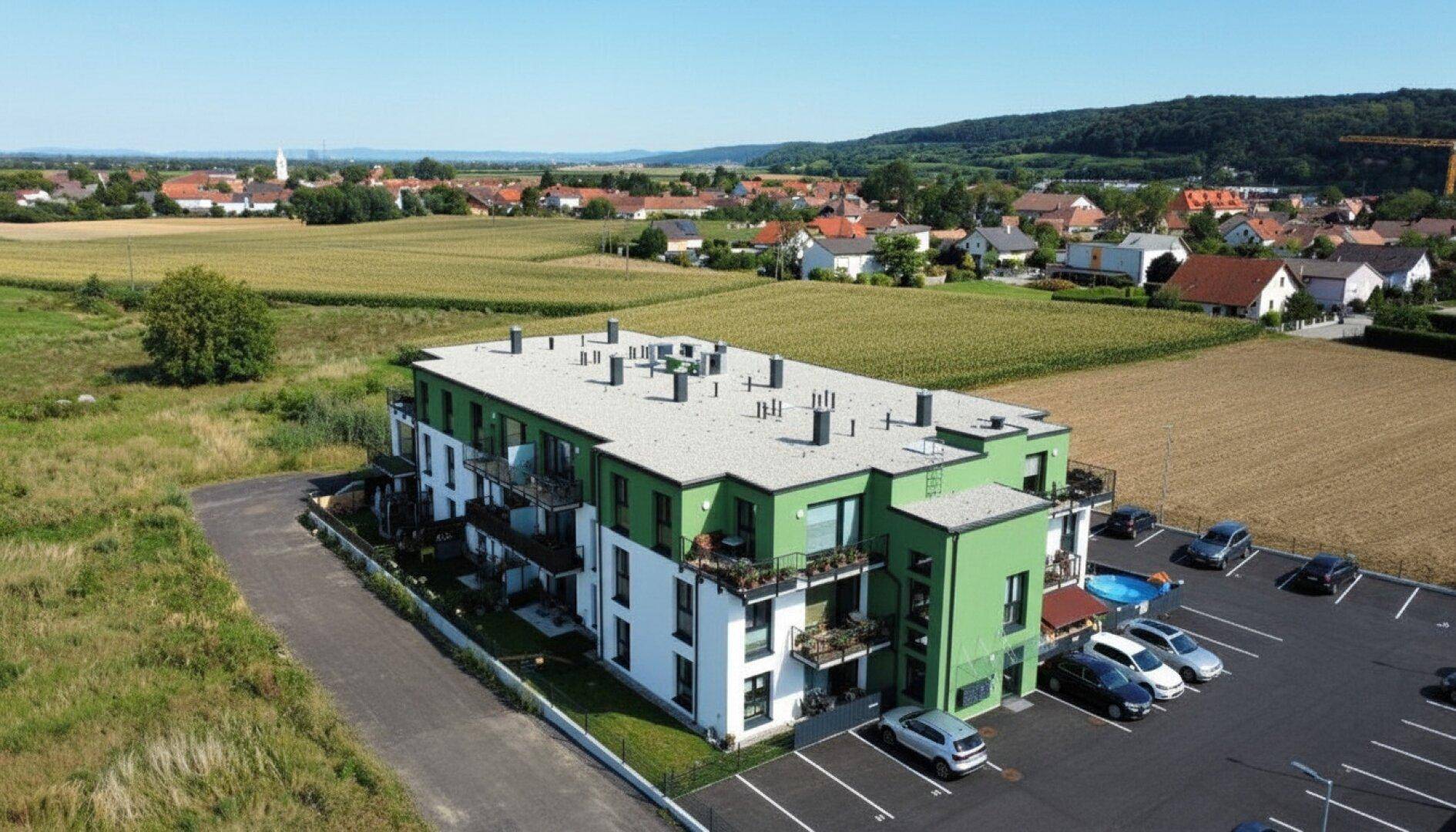 Luftaufnahme des modernen Wohnkomplexes mit Flachdach, umgeben von Feldern und einem Dorf in der Ferne.