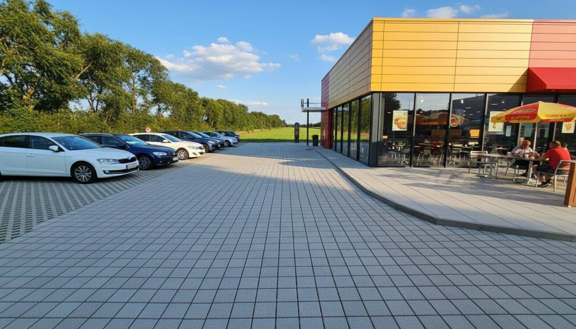 Gewerbegebäude mit gelb-roter Fassade, großem gepflasterten Parkplatz und angrenzender Naturlandschaft.