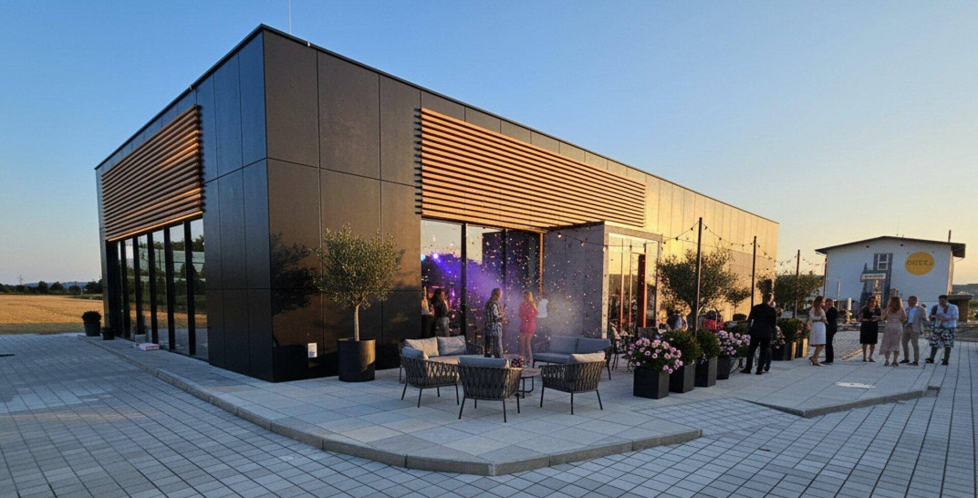 Abendliche Ansicht einer modernen Gewerbeimmobilie mit beleuchteter Terrasse und Gästen im Freien.