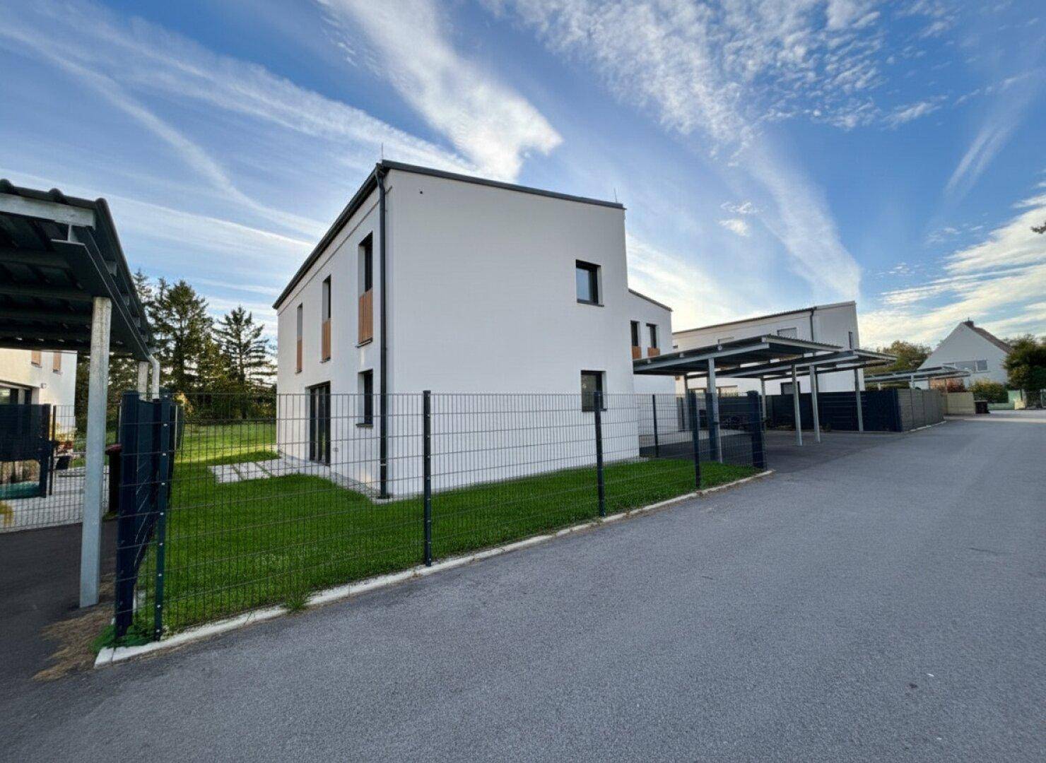Neubau-Reihenhaus mit weißer Fassade, kleinem Garten und Carport im Hintergrund.