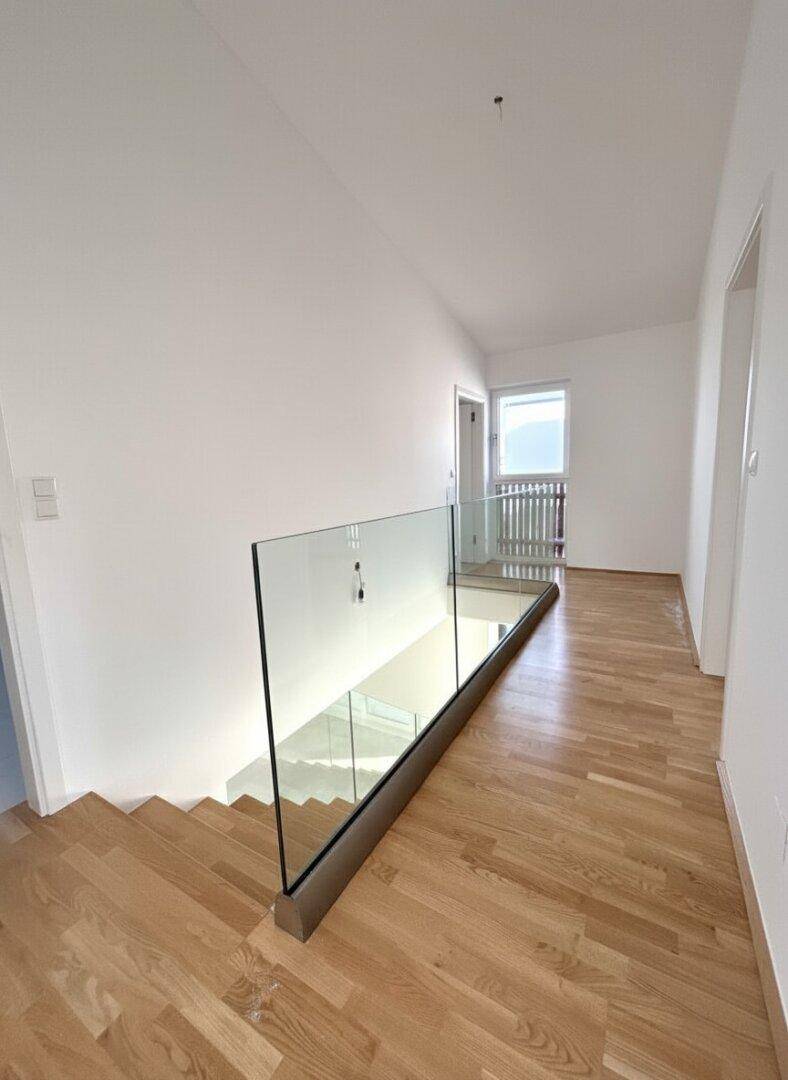 Heller Flur im Obergeschoss mit Holzparkett und modernem Glasgeländer zur Treppe.