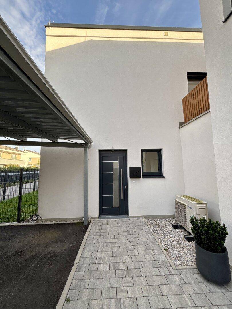 Eingangsbereich eines modernen Hauses mit Carport, gepflastertem Weg und Wärmepumpe.