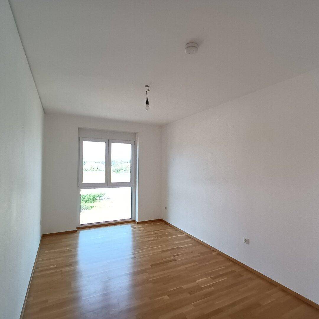 Leeres Schlafzimmer mit Holzboden und einem Fenster, das den Raum hell erscheinen lässt.