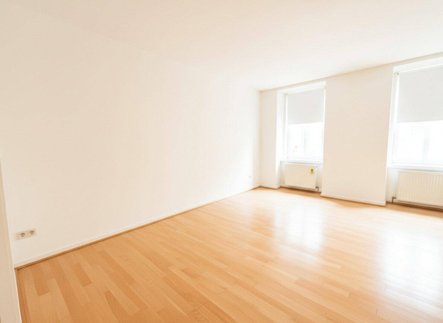 Geräumiges Zimmer mit hellem Holzboden und zwei Fenstern, bereit zur individuellen Gestaltung.