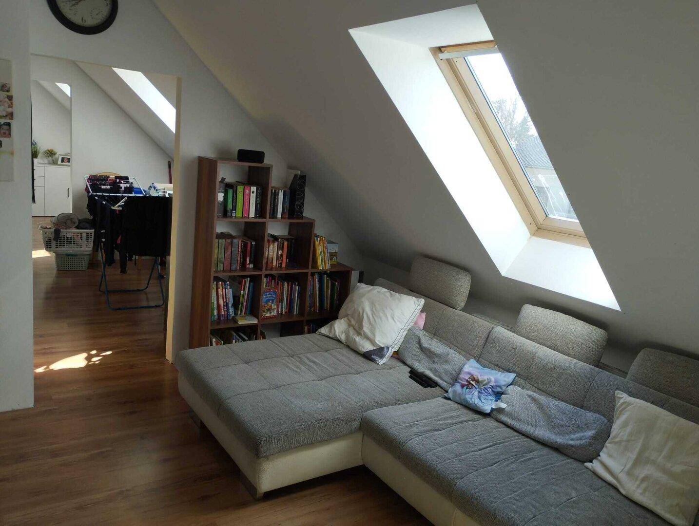 Gemütliches Dachgeschoss-Wohnzimmer mit großem Sofa und Bücherregal unter dem Dachfenster.