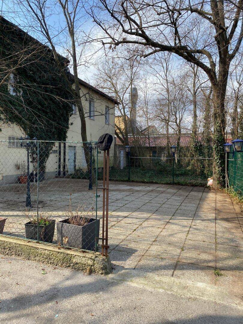 Gepflasterter Außenbereich hinter dem Gebäude mit Bäumen und einem Zaun, der auf sonniges Wetter hindeutet.